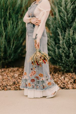 Person wearing a flowing floral maxi dress with pink sleeves holds dried wildflower bouquet against evergreen background.