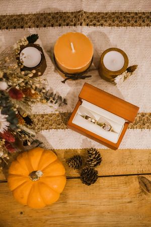Autumn styled flatlay featuring orange pumpkin, pine cones, jewelry box with ring, and candles on wooden surface.