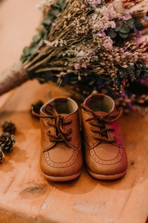Small brown leather baby shoes displayed with dried flowers and pinecones on wooden surface.