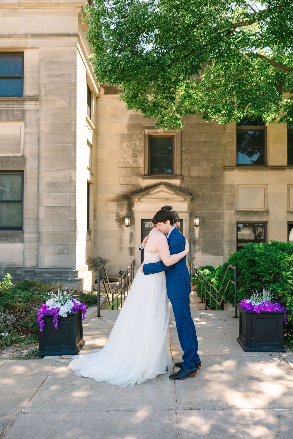 A couple in wedding attire hug in front of First United Methodist Church surrounded by spring greenery and purple flowers in pots.