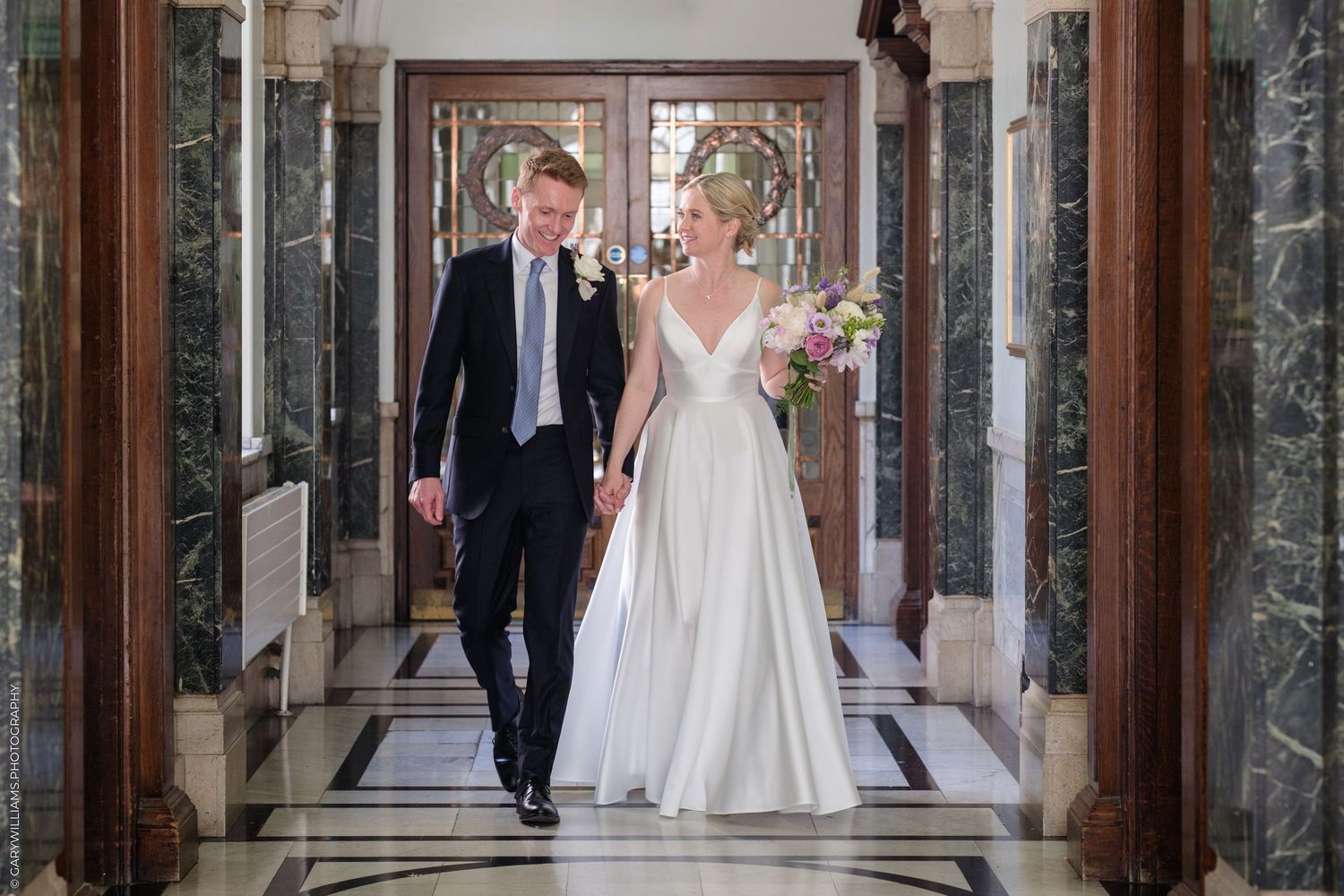 A couple in formal wedding attire walk down an elegant marble hallway with ornate architectural details.