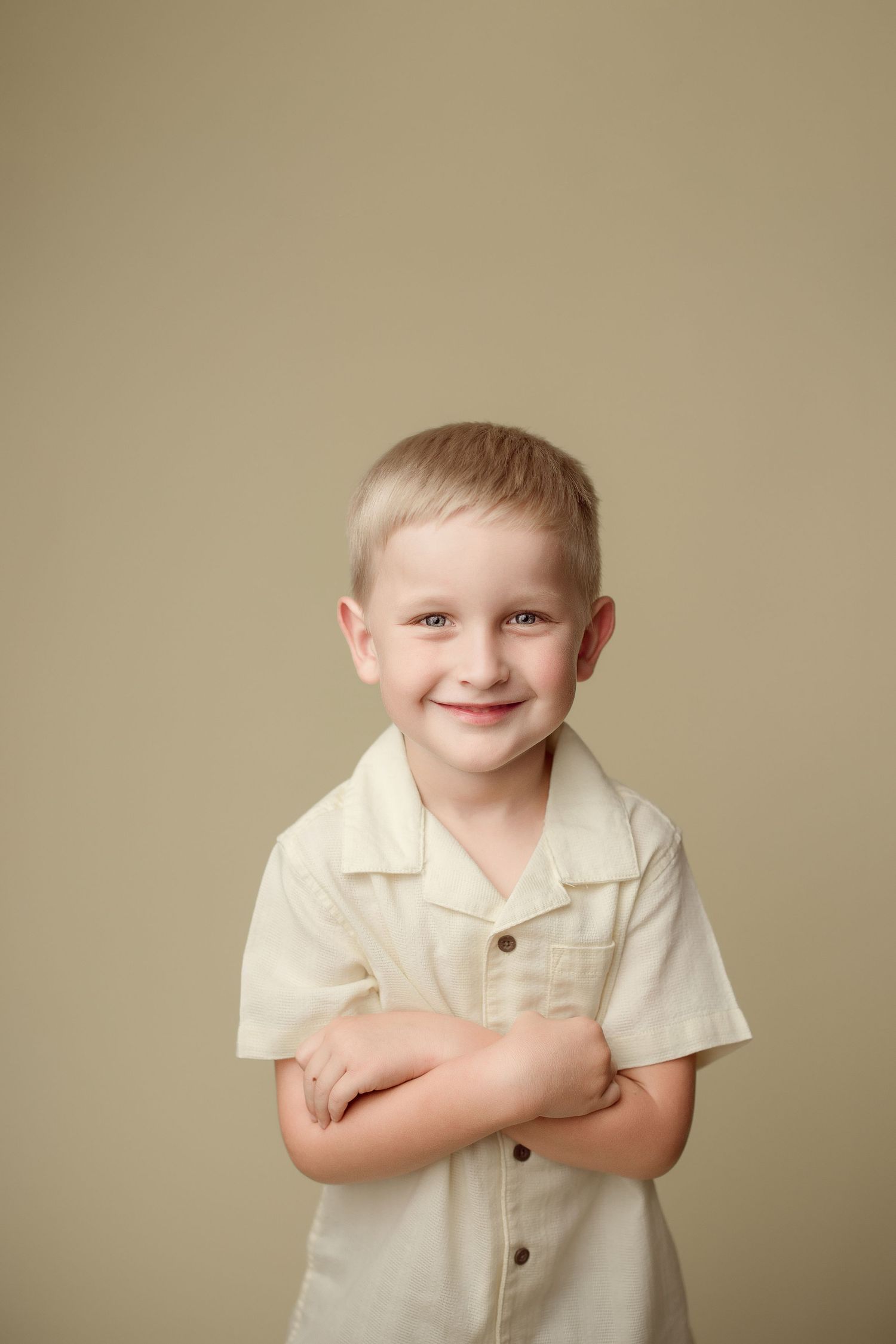 Young child in cream-colored button-up shirt stands with arms crossed against neutral beige background.