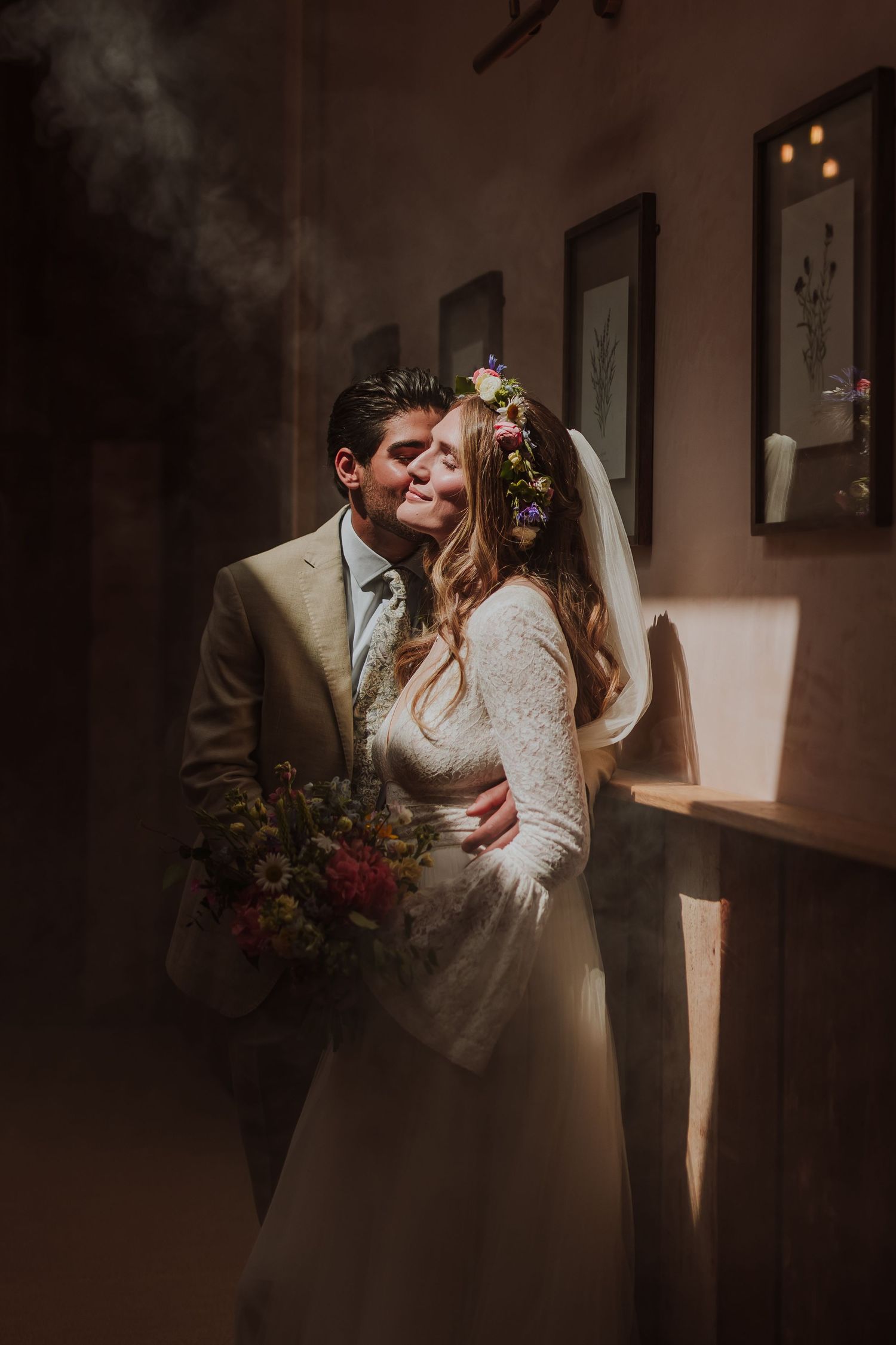 A romantic wedding couple shares intimate moments in a dimly lit indoor setting with moody atmospheric lighting.