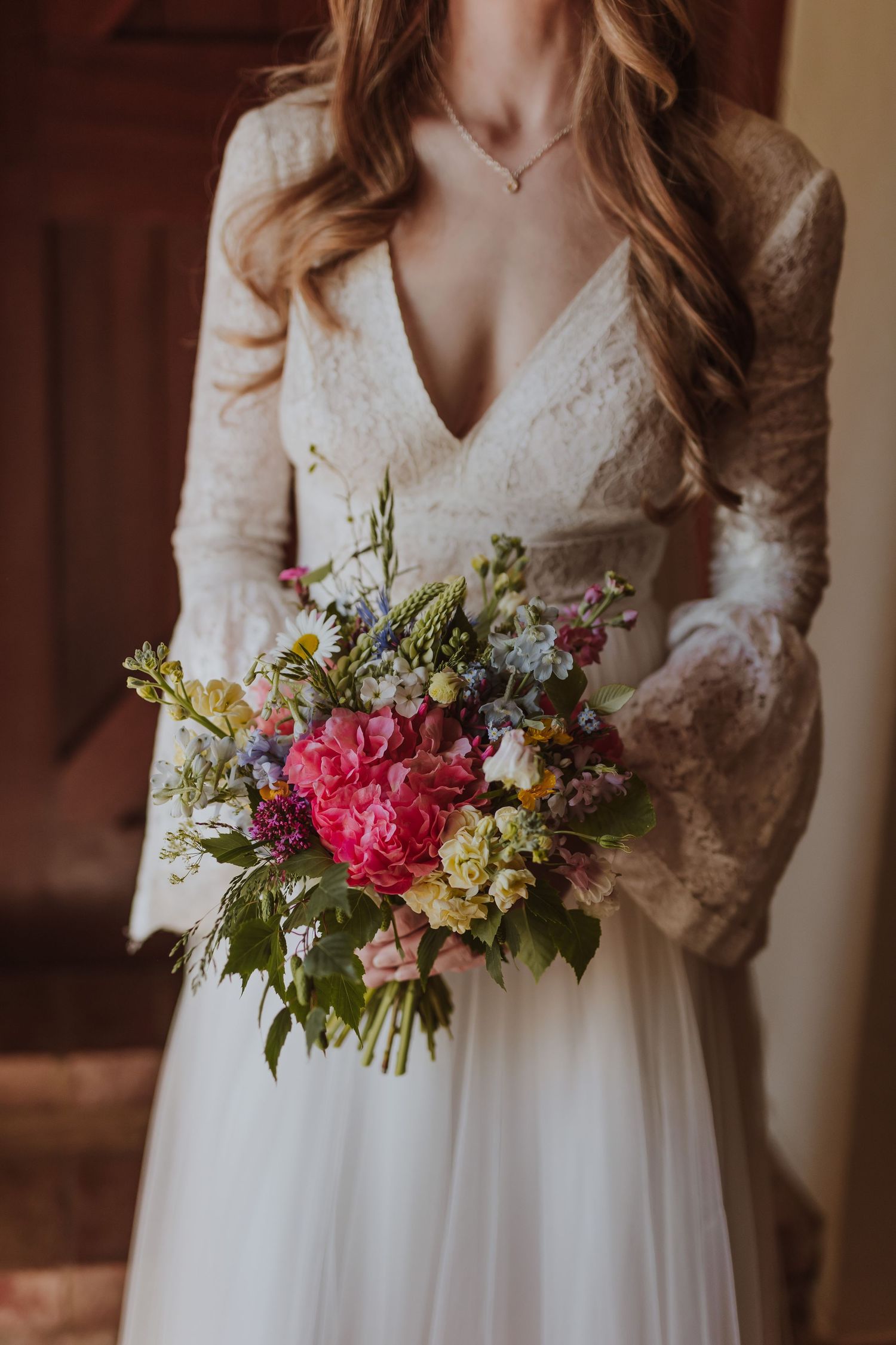 Close-up view of a vintage-inspired bridal gown with lace sleeves and a colourful floral bouquet featuring pink and green blooms.