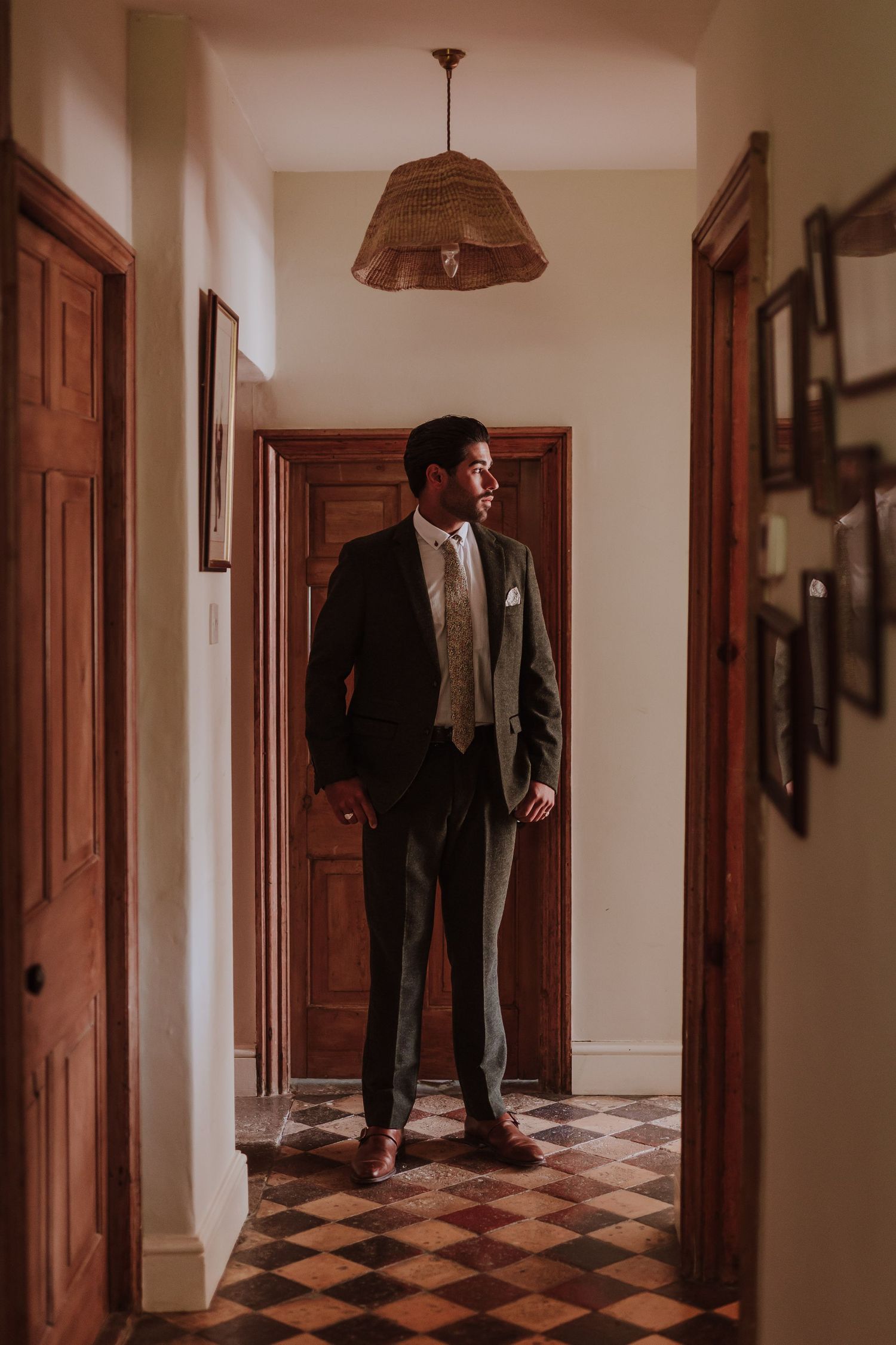 Dark suited groom stands in a hallway with wooden doors and a checkerboard floor beneath a woven pendant light.