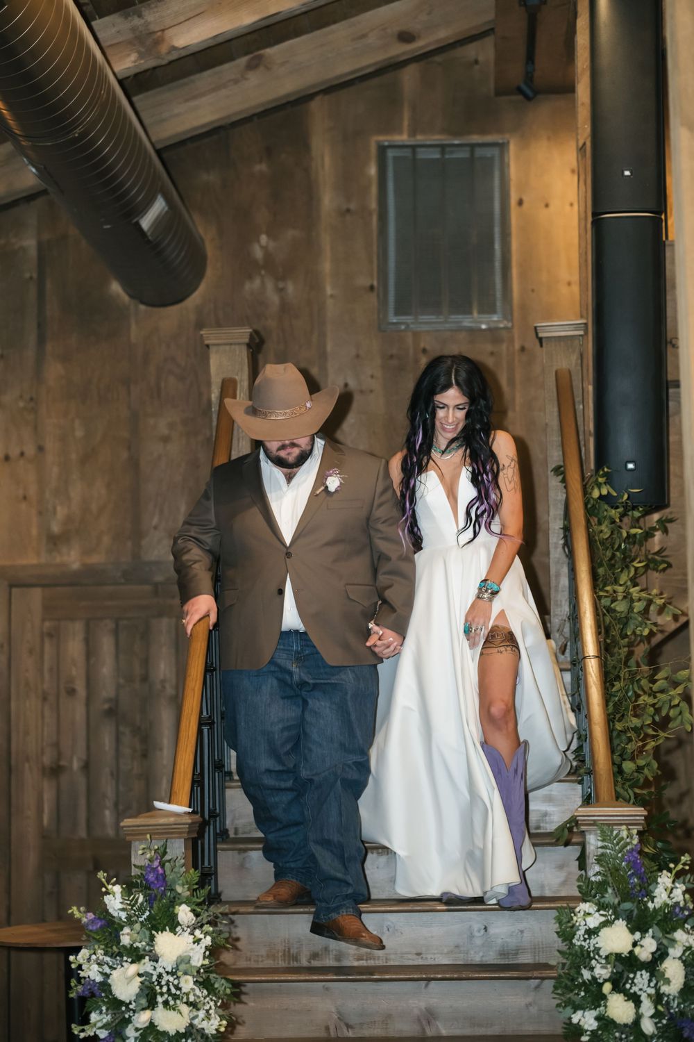 A couple descends rustic wooden stairs decorated with floral arrangements at a barn wedding venue.