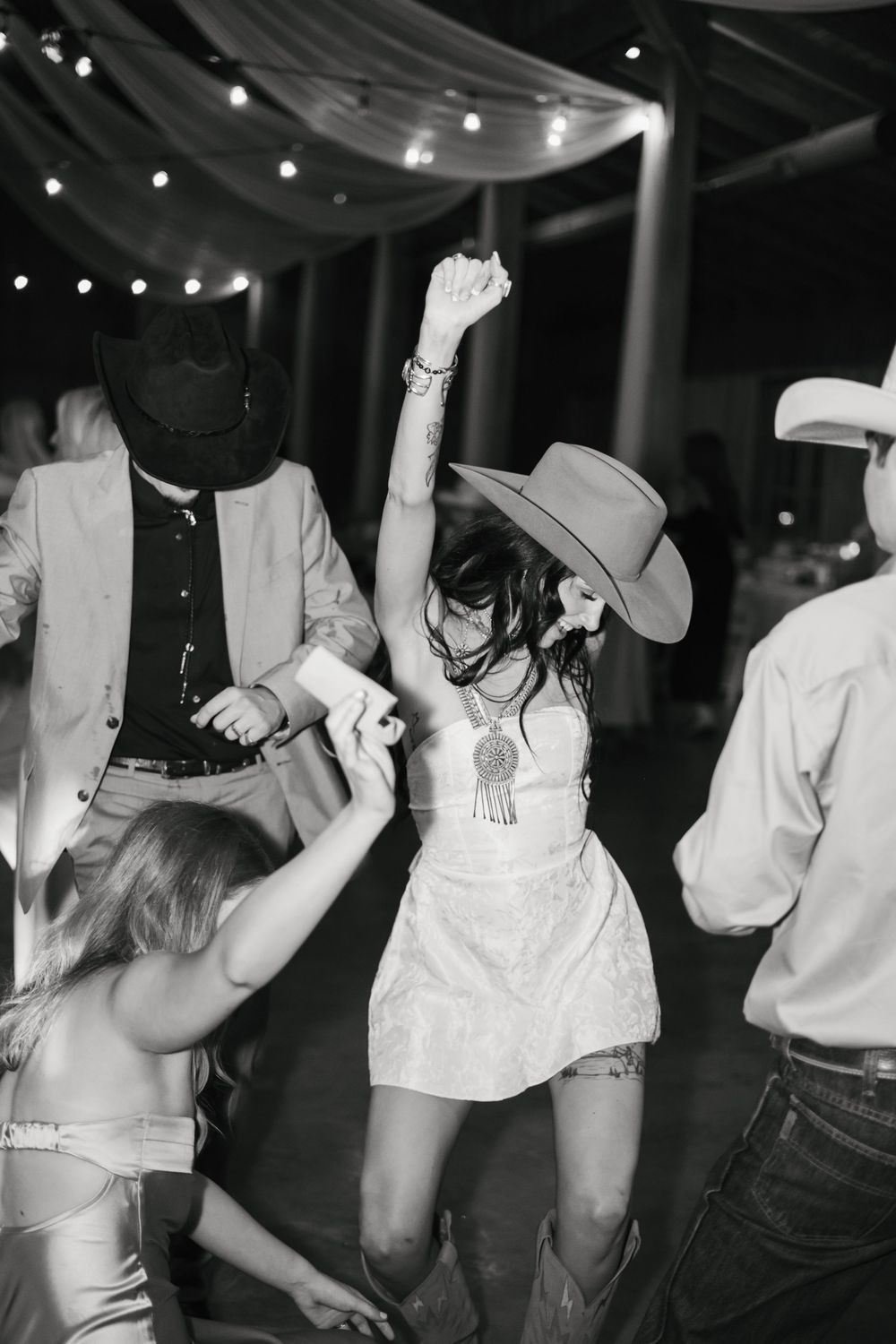 Group of people dancing and having fun at a party under white string lights in a black and white photo.