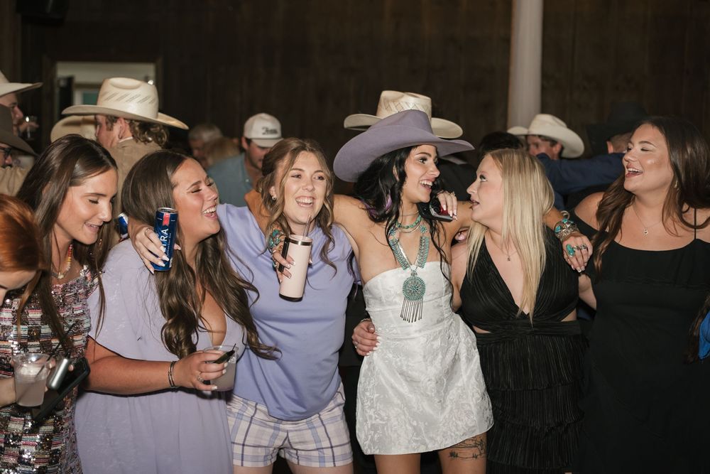 A group of friends wearing western hats and summer dresses laugh together at a party under dim lighting.