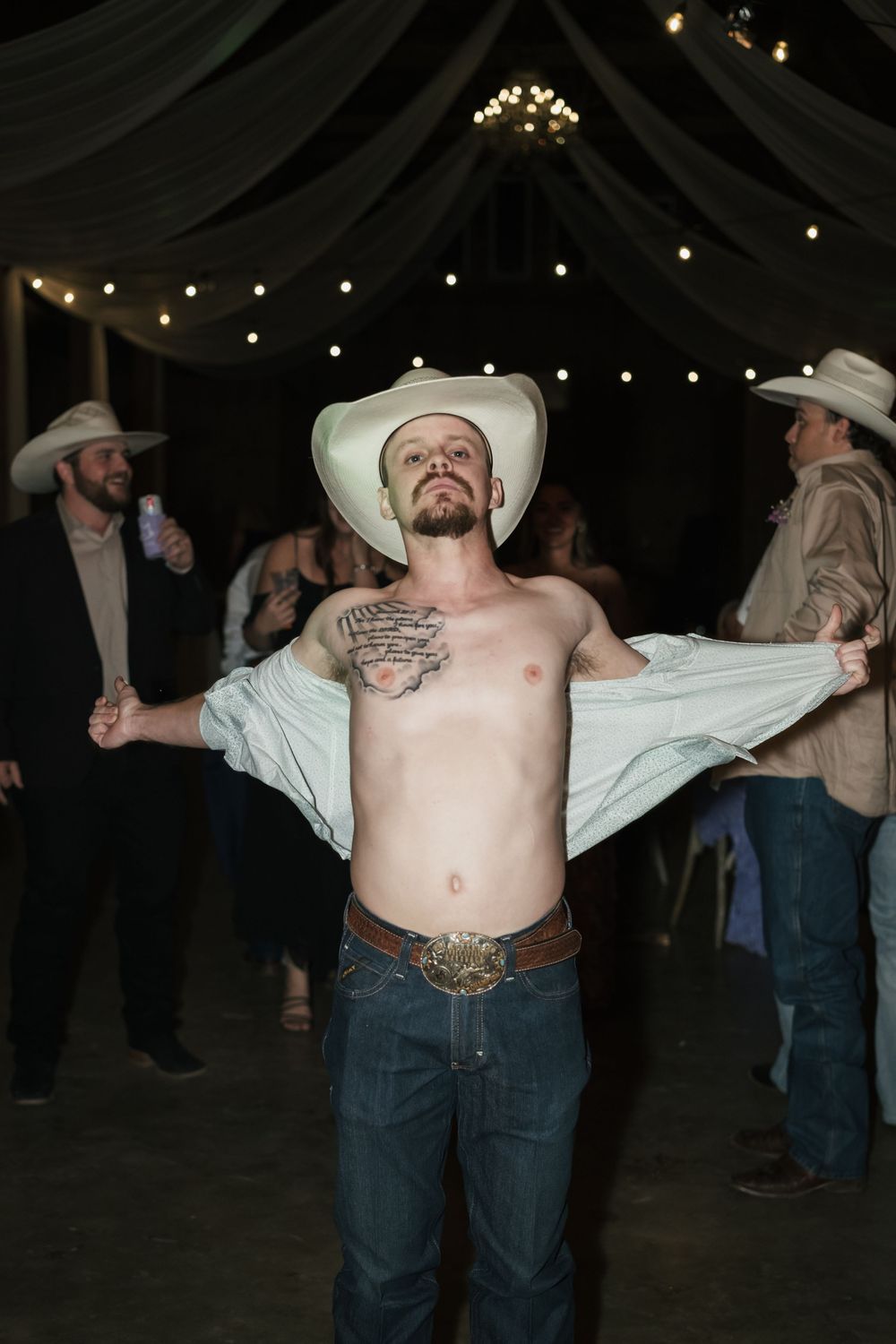 A shirtless man in cowboy hat and jeans strikes a dramatic pose under string lights at an indoor venue.