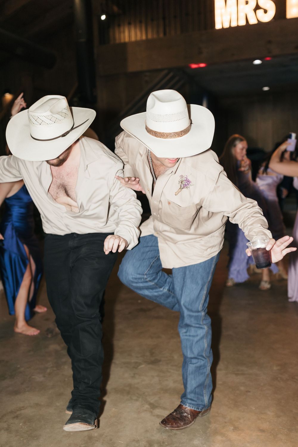 Guests wearing white cowboy hats perform a synchronized line dance on a concrete floor at a western-themed event.