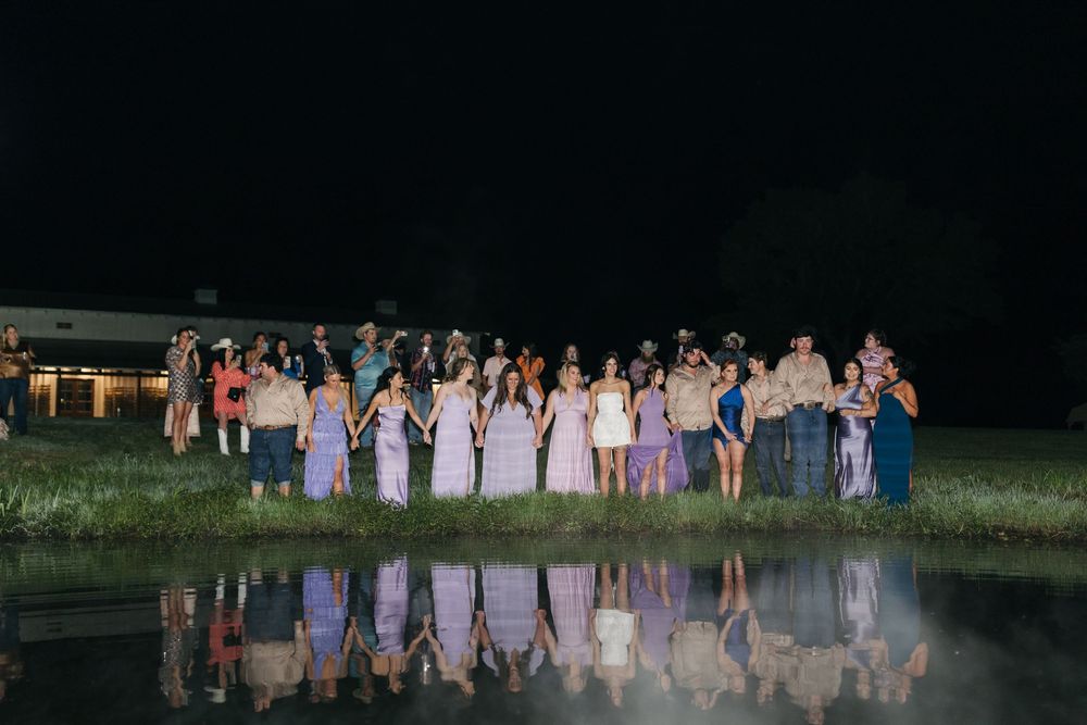 Wedding party members stand at night beside a reflective pool creating a mirror-like image in the water.