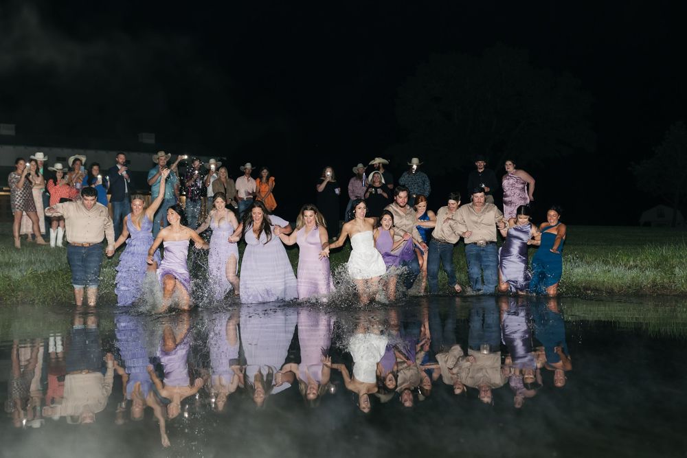 Wedding party runs together forming a cheerful group photo at dusk in an outdoor garden setting.