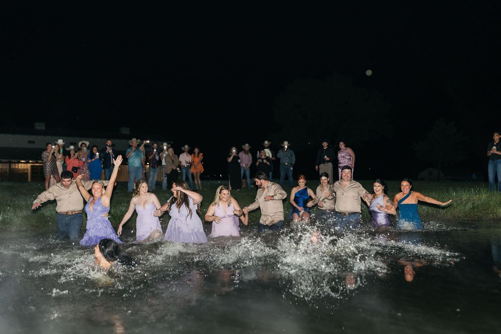 A large group of wedding guests create a water splash while jumping into a lake at night.