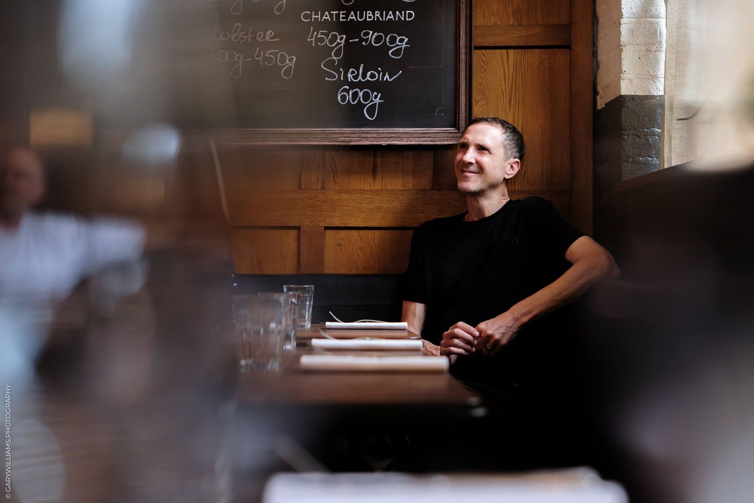 Someone wearing black clothing sits at a wooden table near a chalkboard menu in a dimly lit restaurant setting.