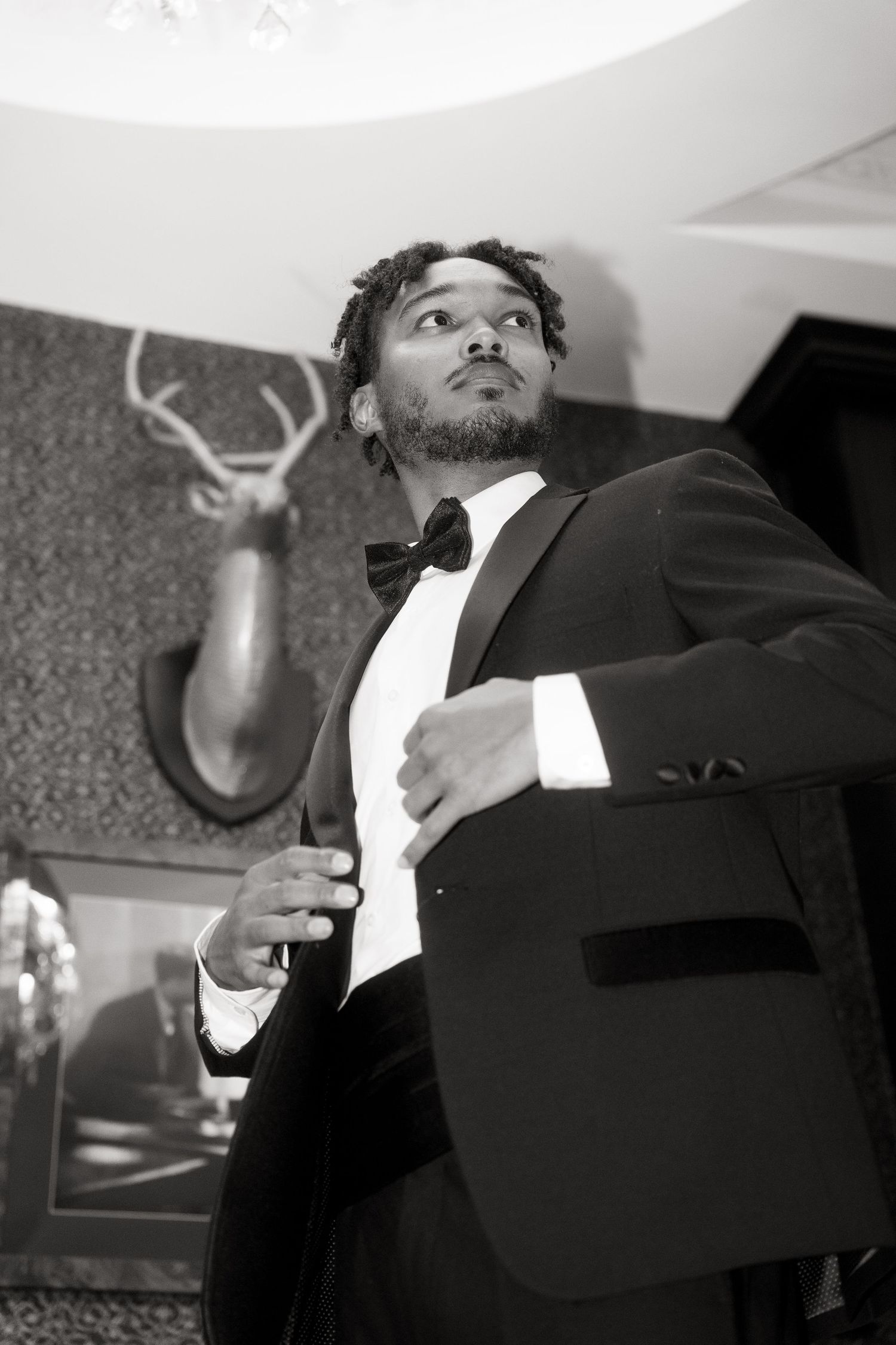 A person in a formal black tuxedo and bow tie poses confidently at a low angle with mounted antlers visible in background.