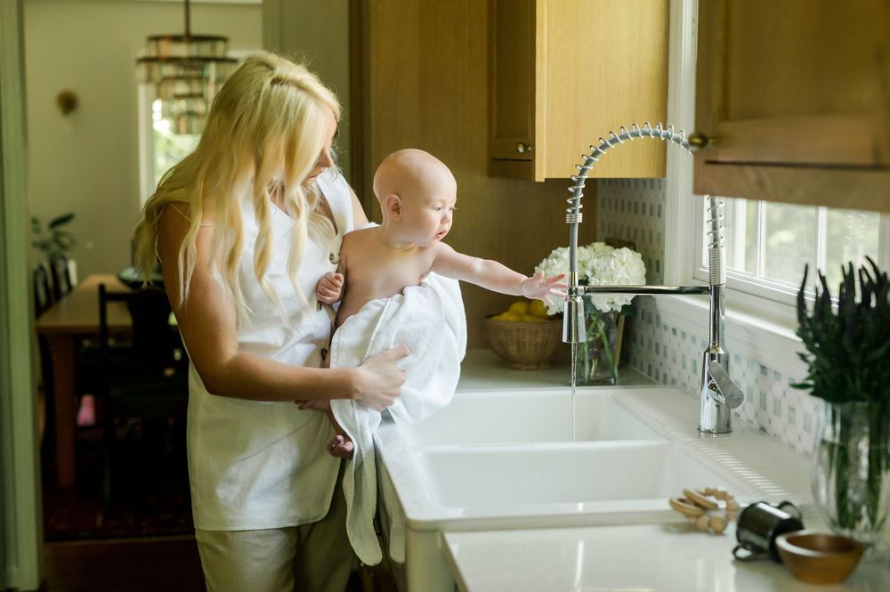 A parent holds a baby reaching toward a modern kitchen faucet at a white farmhouse sink in a sunny room.