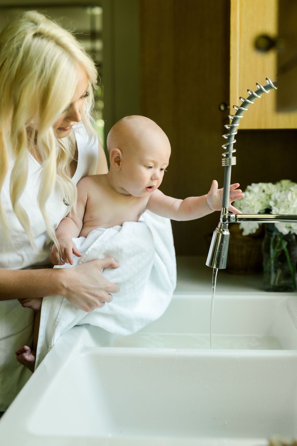 A mother helps her baby play in white bathtub with handheld faucet during bathtime.