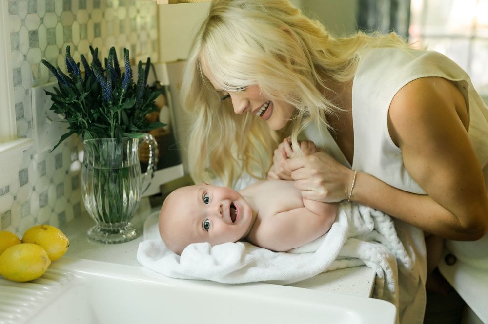 A parent leans over a smiling baby lying on a changing table next to a vase of flowers and lemons on the counter.