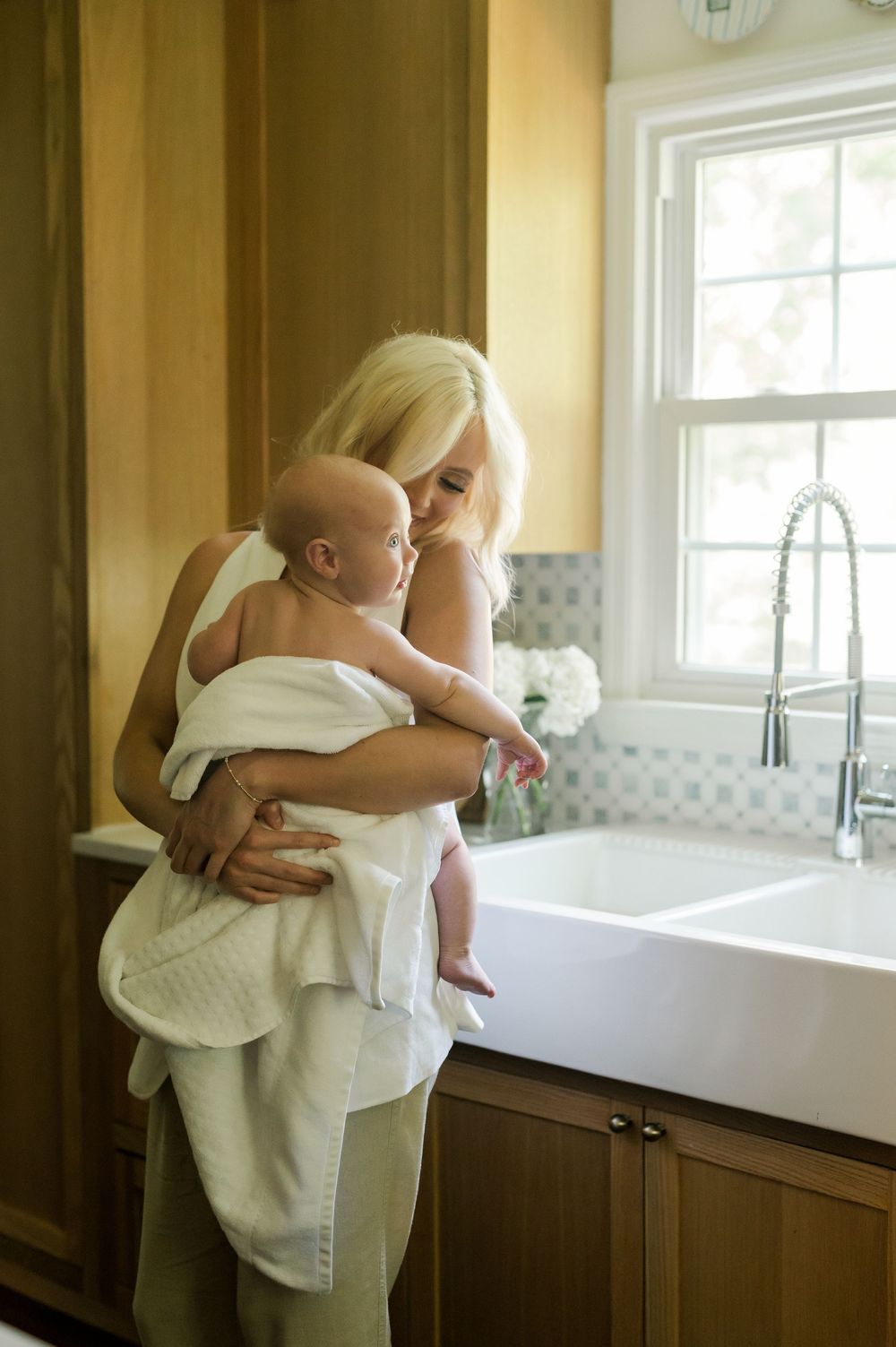 Figure wrapped in white towel holding baby near farmhouse style sink in bright kitchen with yellow walls.