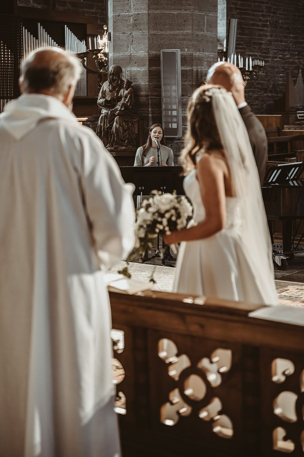 Vigeslakt med solosång i Klosterkyrkan Vadstena, brudparet och prästen står vända mot sångerskan. Bröllopsfotograf Josefin Landhagen.