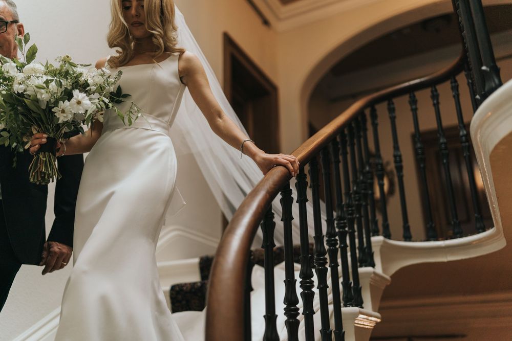 A bride in a sleek white wedding dress ascends an elegant staircase at Cleatham Hall.