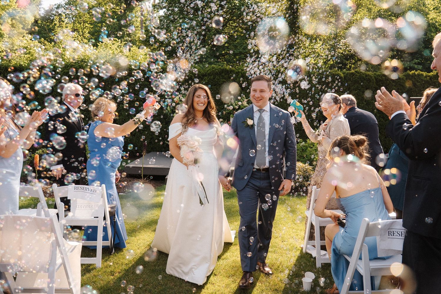 Newlywed couple walks down outdoor garden aisle while guests throw white flower petals during sunny wedding ceremony.