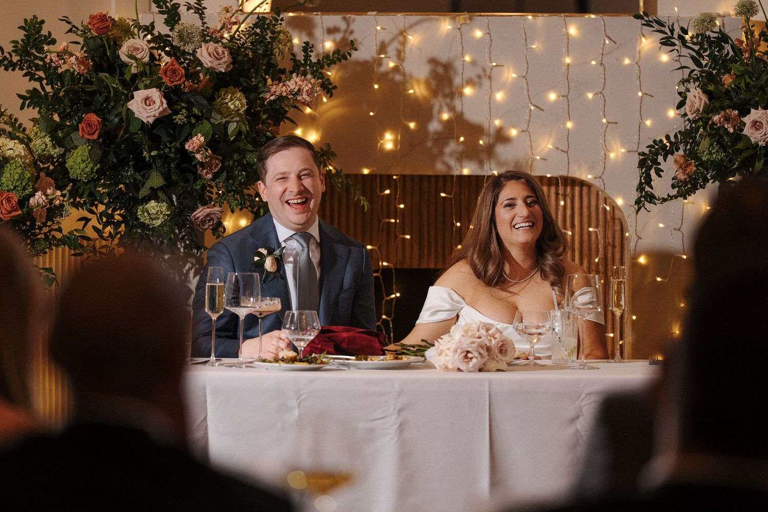 Wedding reception guests share laughter and conversation at elegant tables decorated with twinkle lights and floral arrangements.