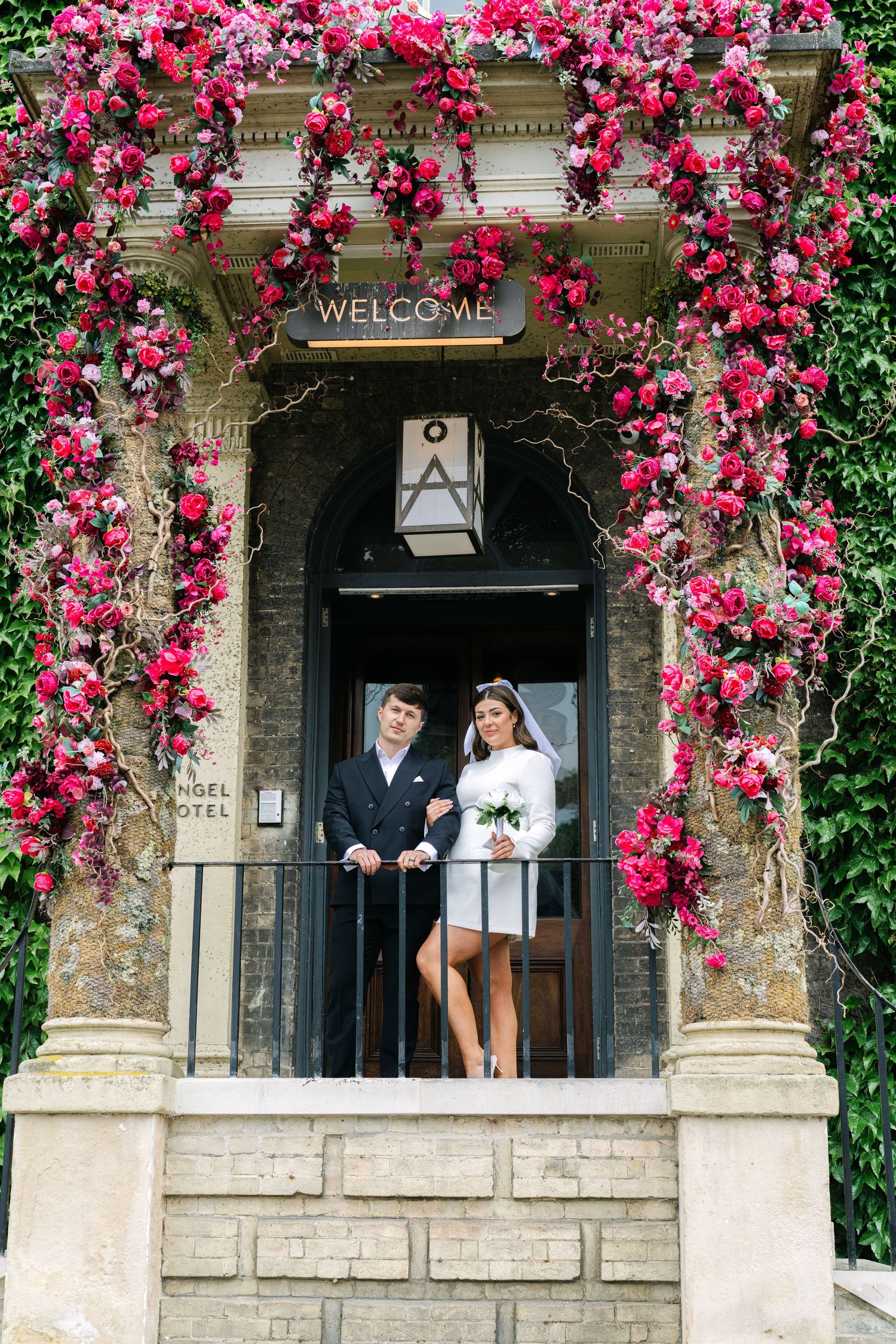 A wedding couple stands in an entryway adorned with vibrant pink climbing roses and a welcome sign.