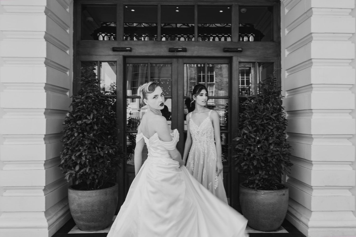 Two brides in elegant wedding gowns share a moment together in a doorway framed by large potted plants in black and white.