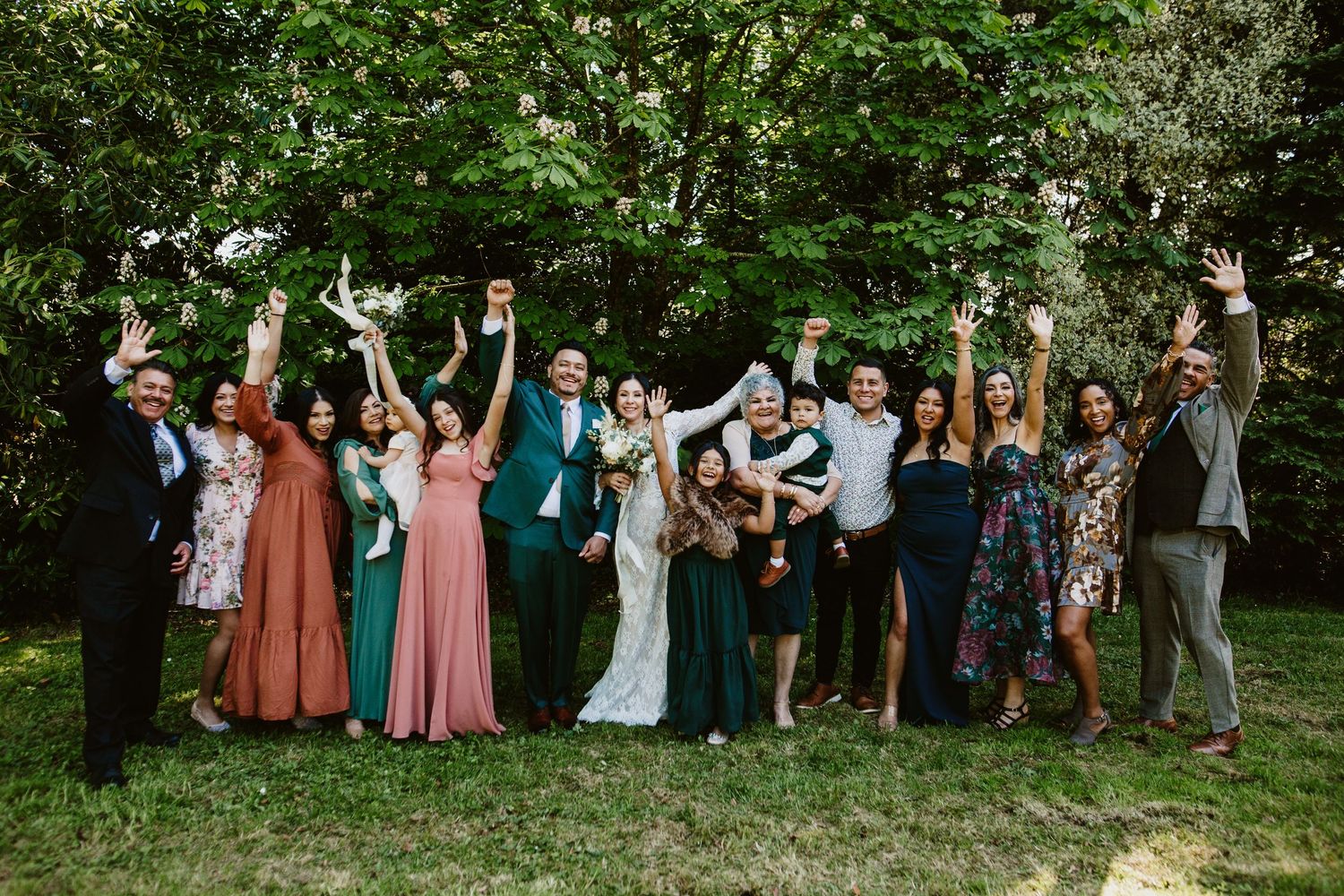 Wedding party stands together outdoors raising their arms joyfully in celebration against green foliage background.