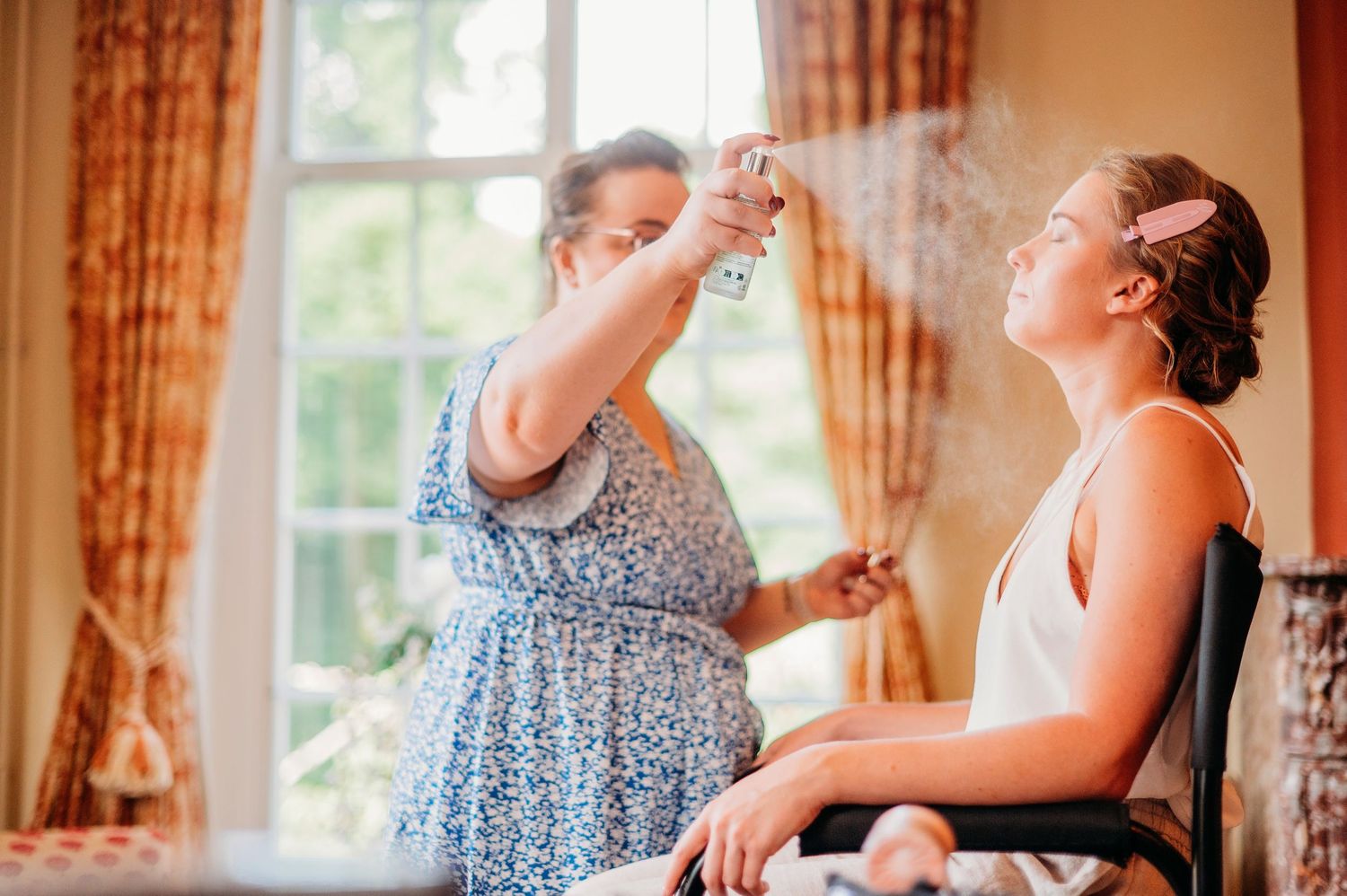 A hairstylist in a floral dress styles bride's hair in sunlit room with elegant curtains and window backdrop.