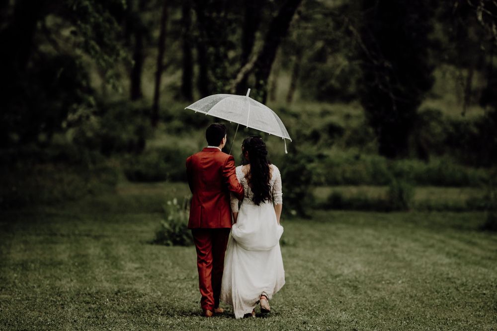 A couple in red suit and white dress share an umbrella while walking together on a lush green lawn.