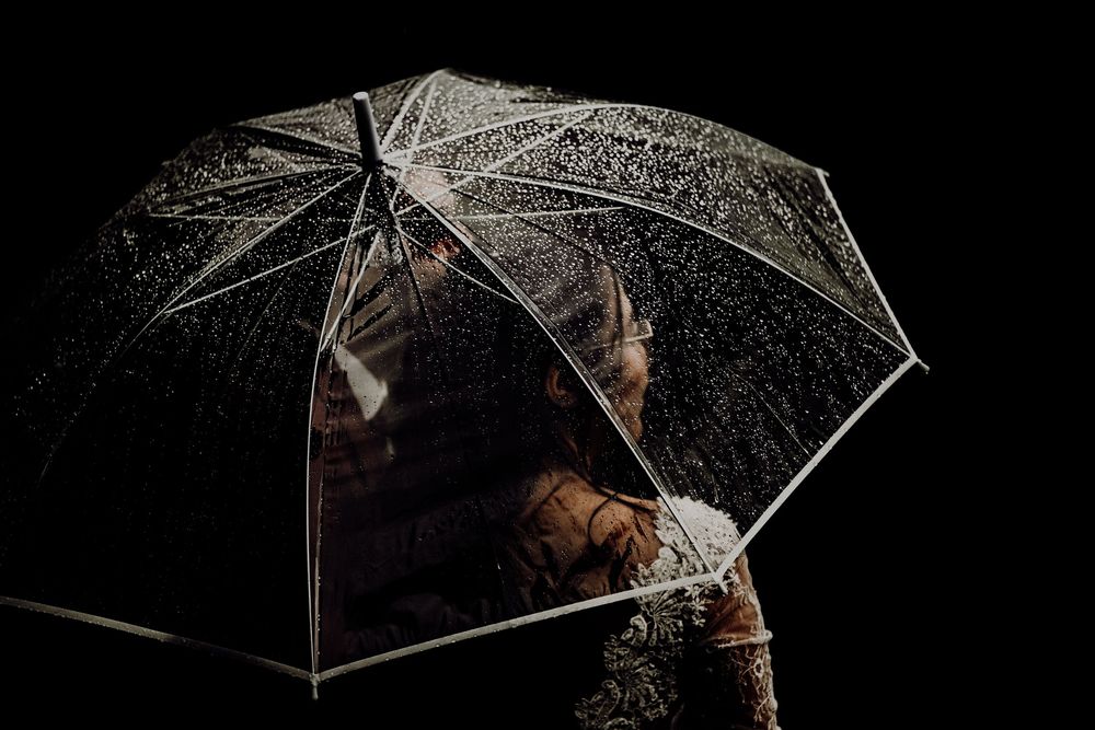A person holds a clear umbrella while water droplets glisten against a dark background in dramatic lighting.