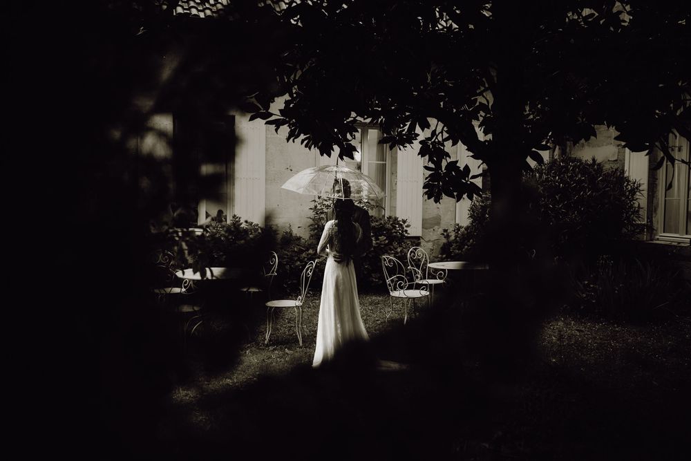 A solitary figure in a flowing white dress stands in a shadowy garden with a dark, vintage-style aesthetic.
