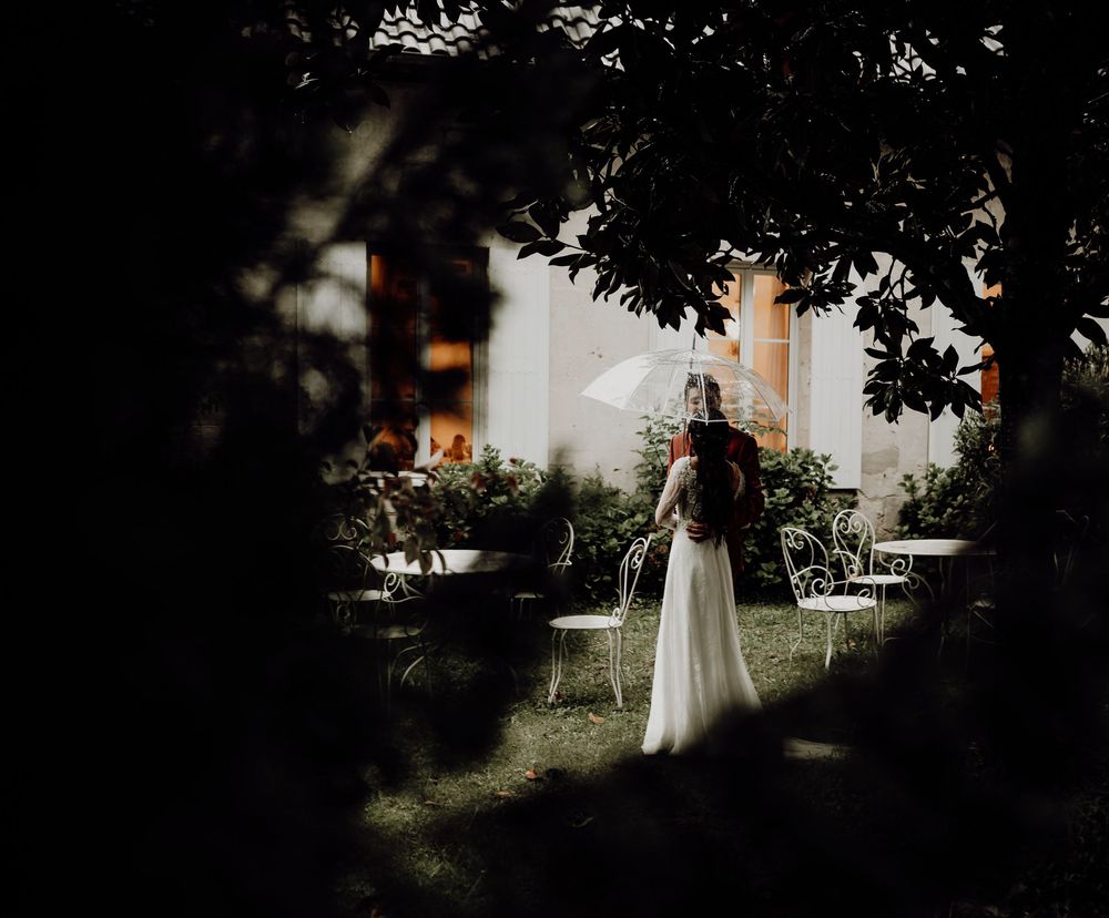 Dark silhouette of a person in a long white dress standing in a garden with trees and sunlight in the background.