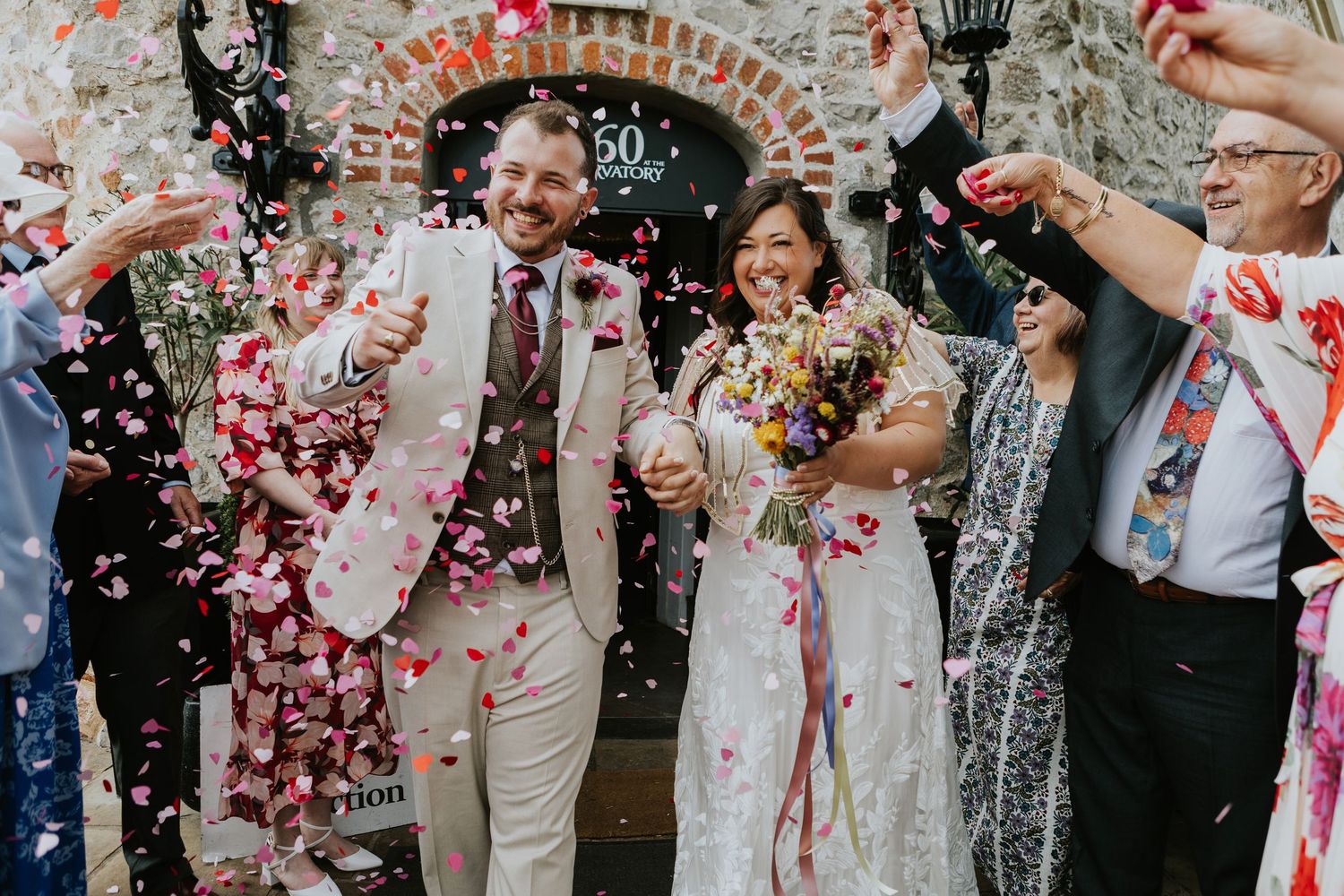Wedding celebration with confetti being thrown over couple as they exit stone archway amid joyful atmosphere.