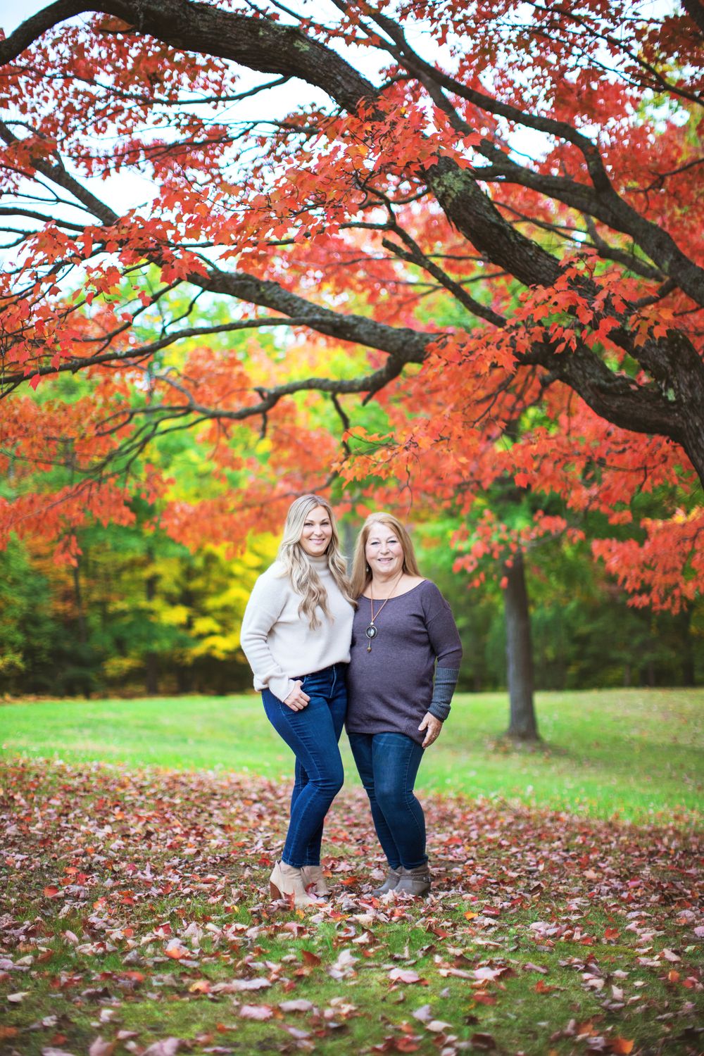 Two people stand together under vibrant red fall maple trees on a grassy lawn with autumn leaves scattered below.