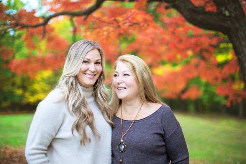 Two women smiling together outdoors with vibrant red autumn foliage in the background.