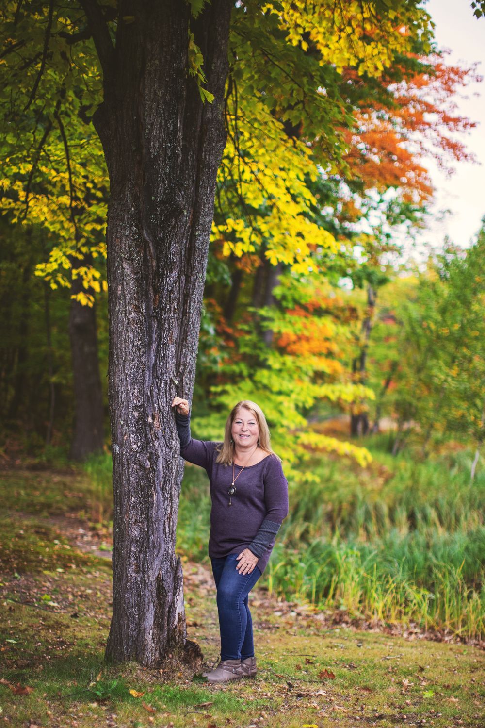 A figure stands beside a large tree trunk against a backdrop of autumn foliage in a woodland setting.