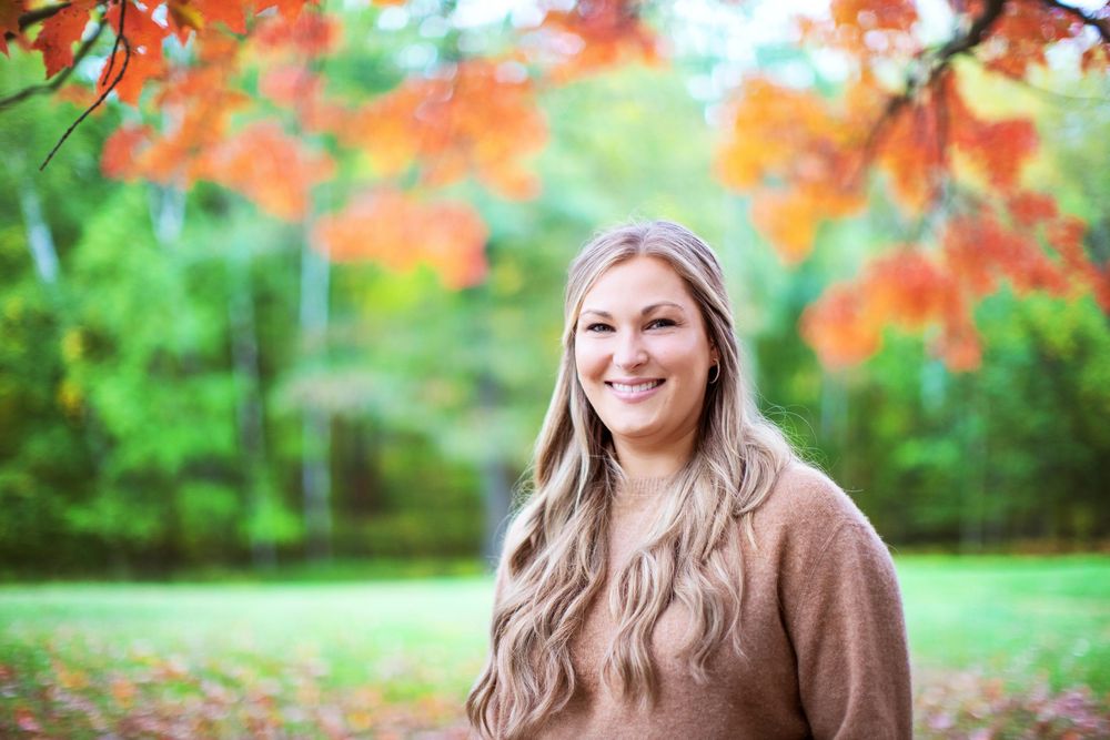 Portrait of person in beige sweater standing beneath bright red autumn foliage in outdoor park setting.