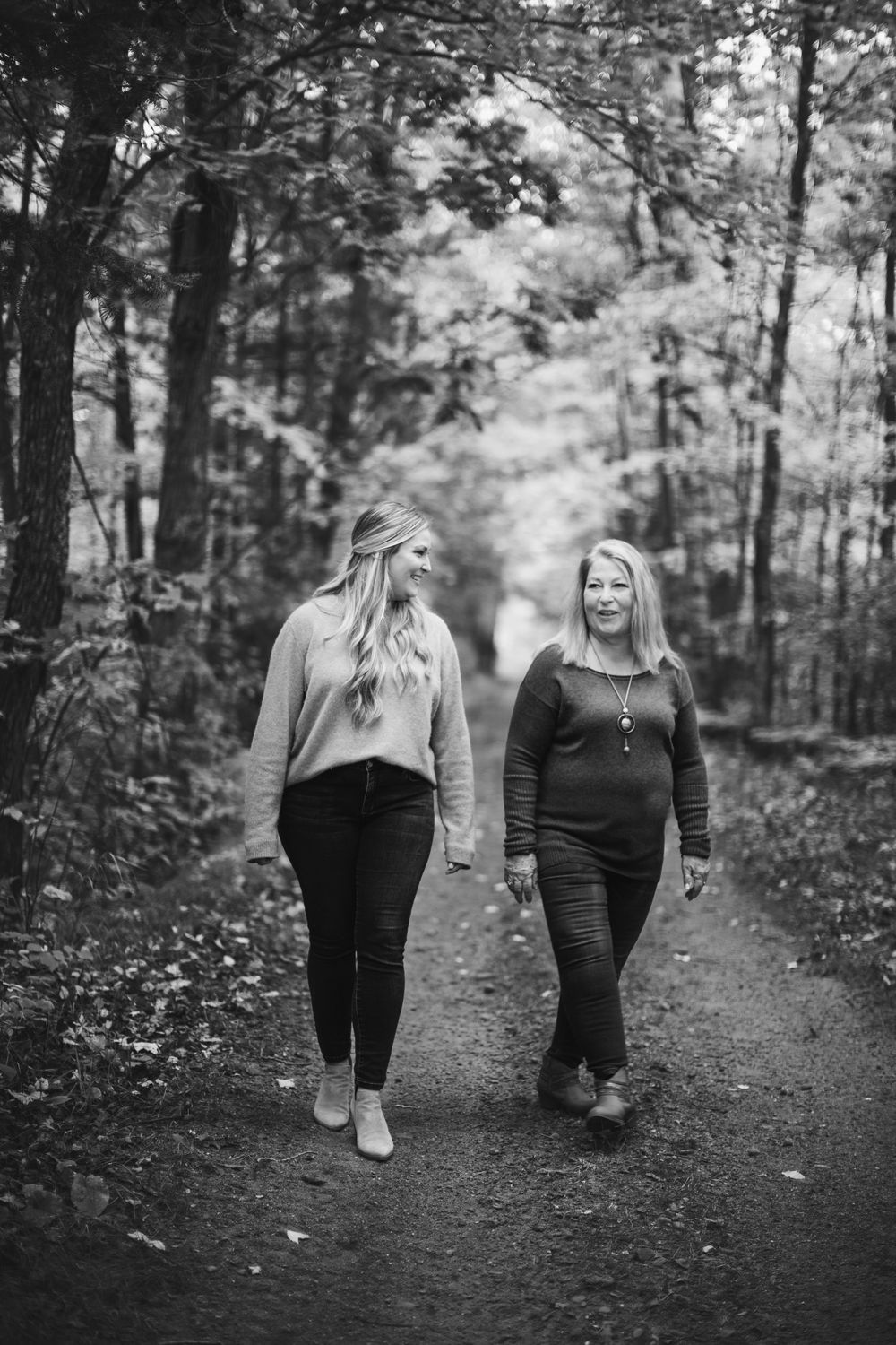 Two people walking together and smiling on a forest path surrounded by tall trees.