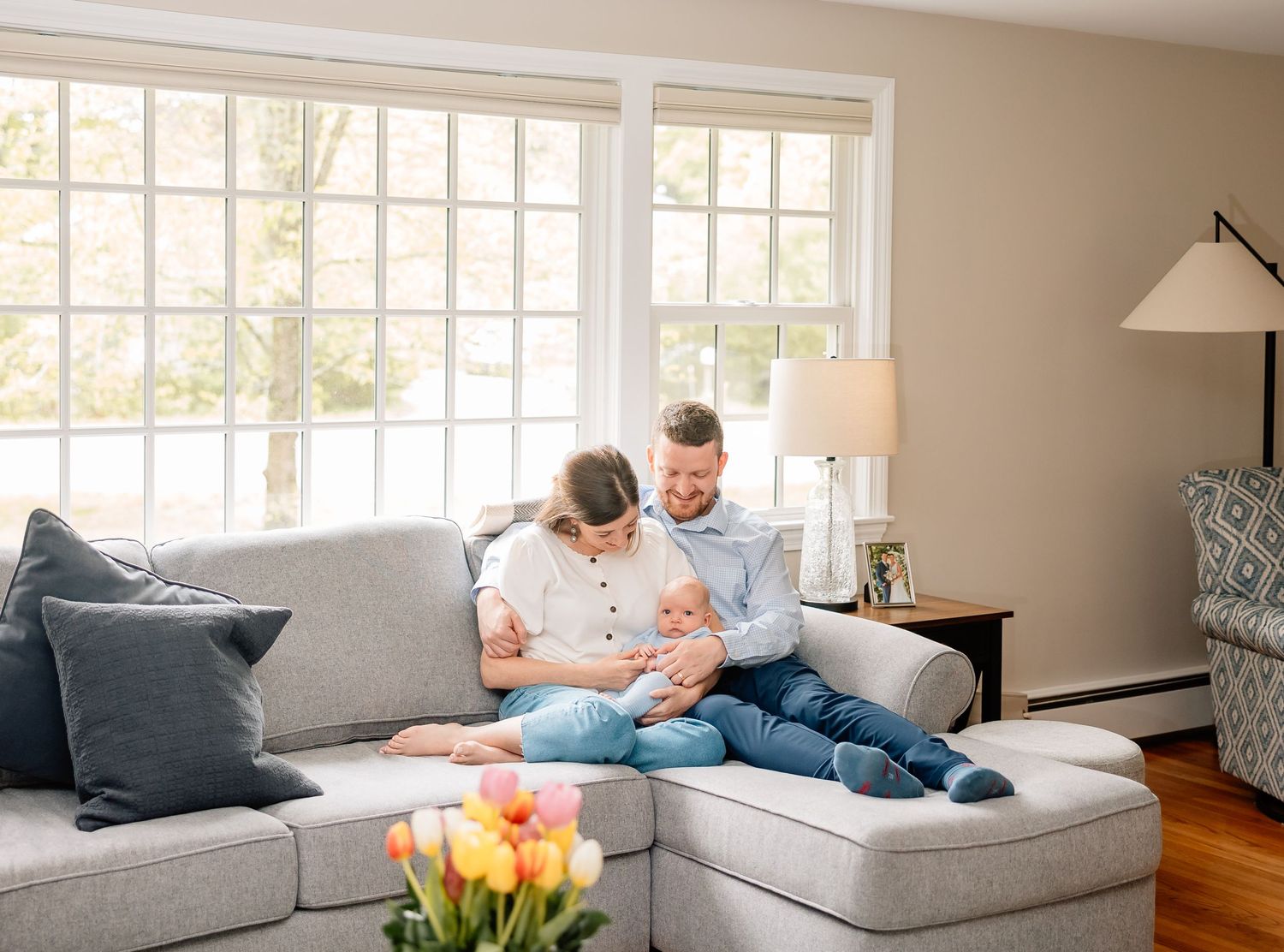 A family relaxes together on a grey sectional sofa in a bright living room with tulips in the foreground.