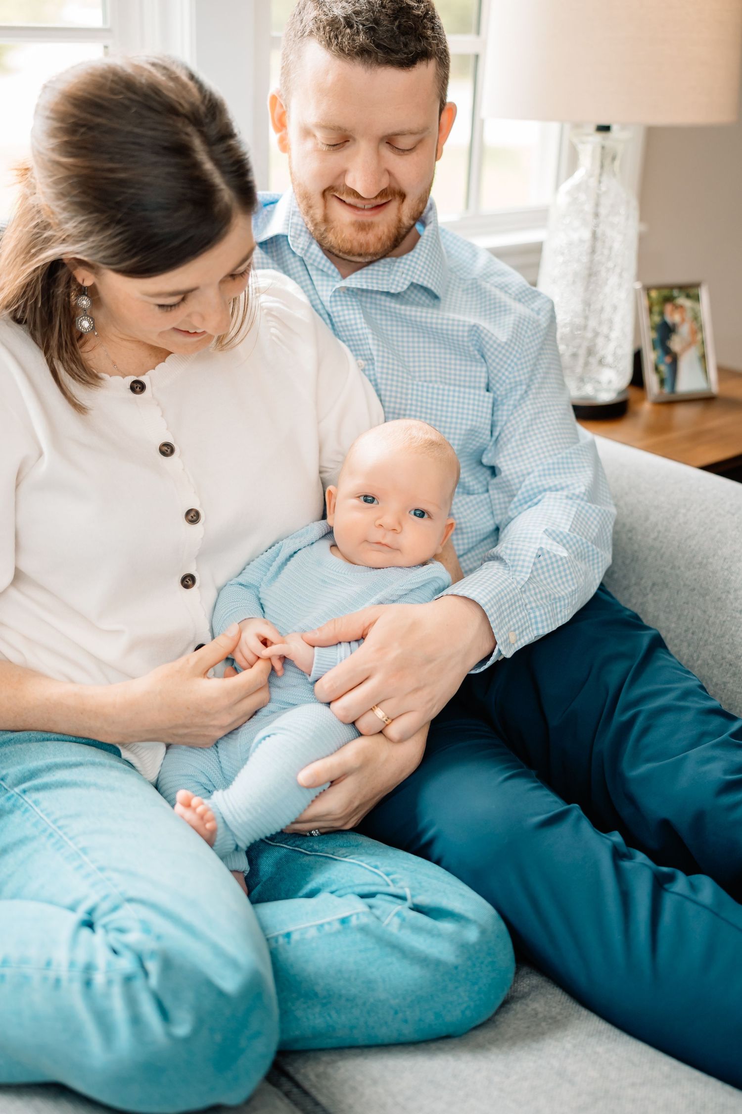 A couple sits on a gray couch cradling their baby in light blue clothing while sharing an intimate family moment.