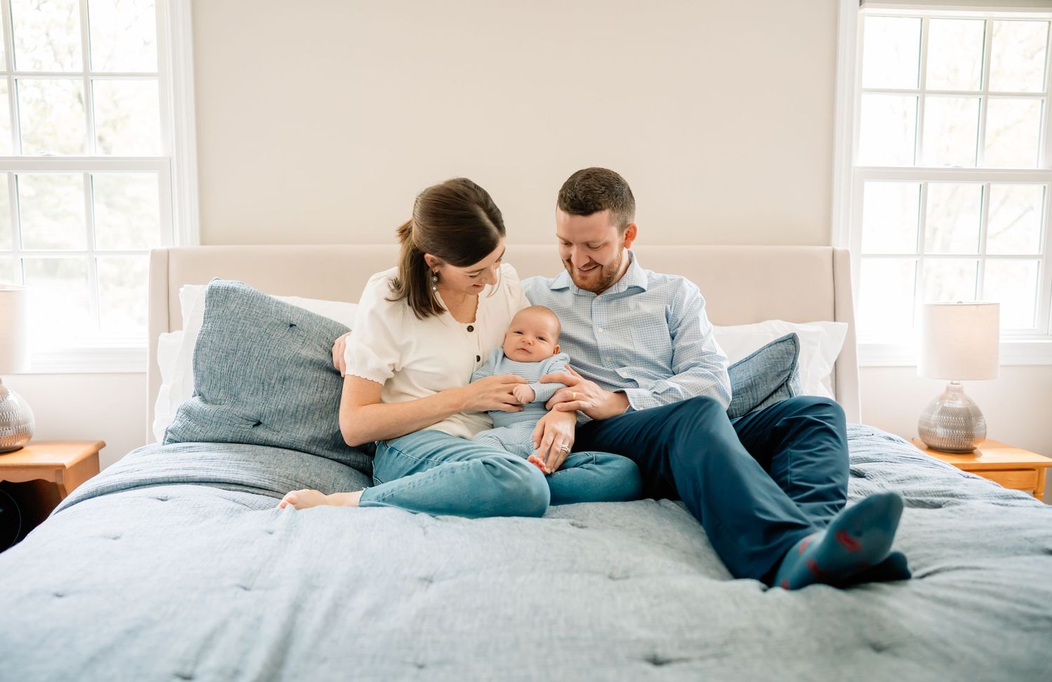 Parents sitting on bed with baby sharing tender family moment together in bright sunlit bedroom.