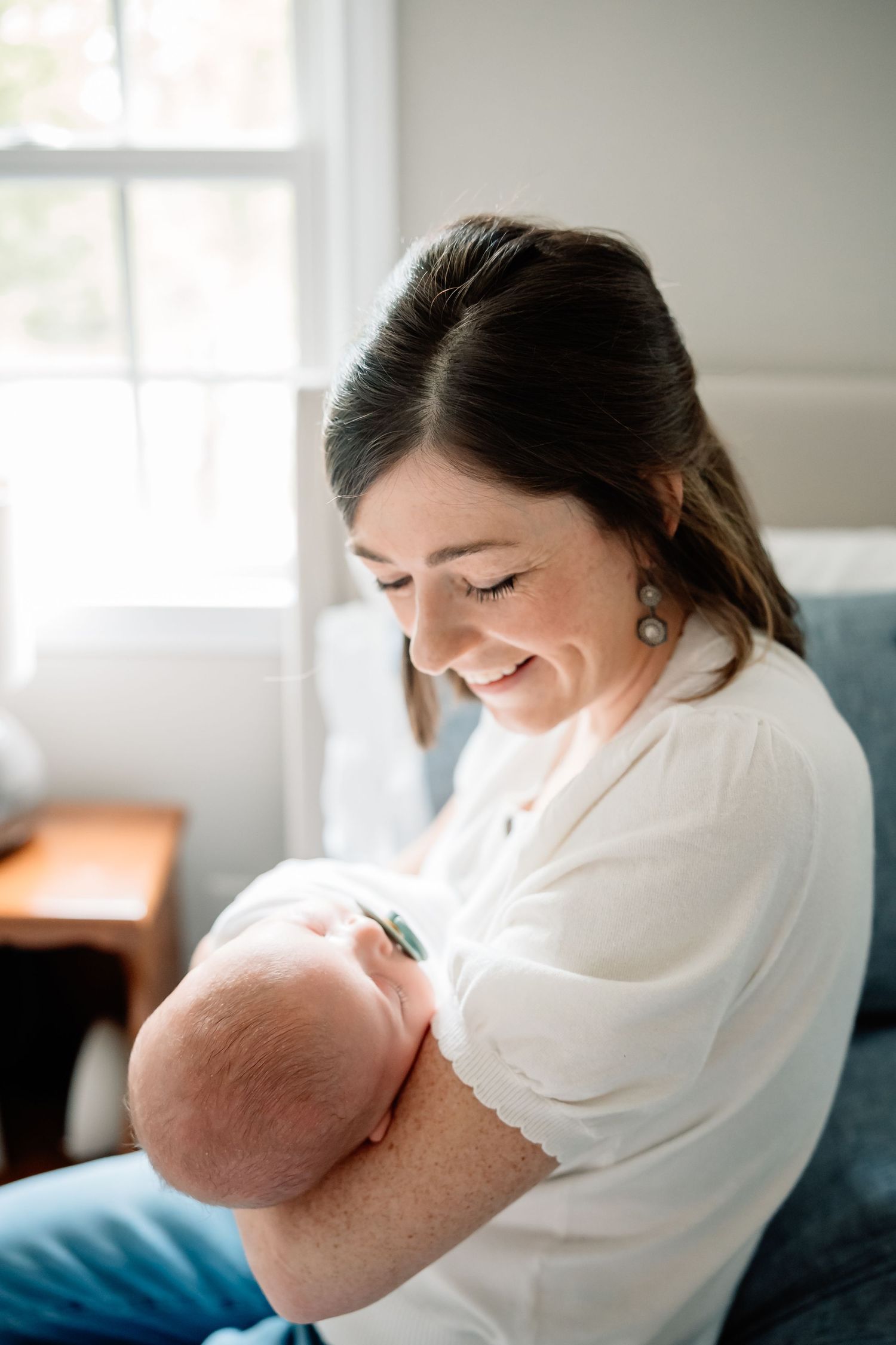 Parent in white shirt sits on blue chair holding and lovingly gazing down at their newborn baby in a bright room.