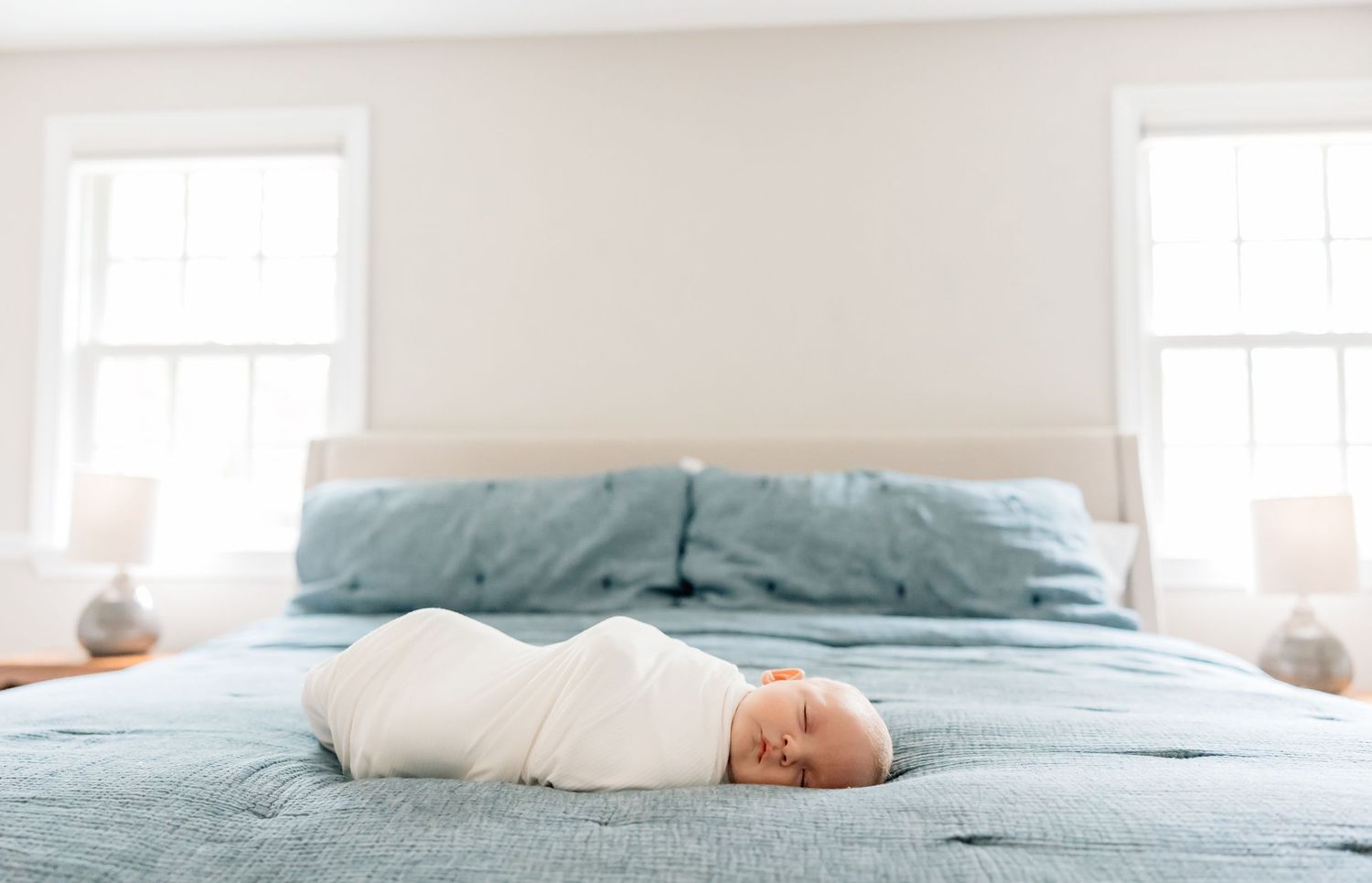 A newborn baby swaddled in white lying at the foot of a bed with blue bedding and bright windows.