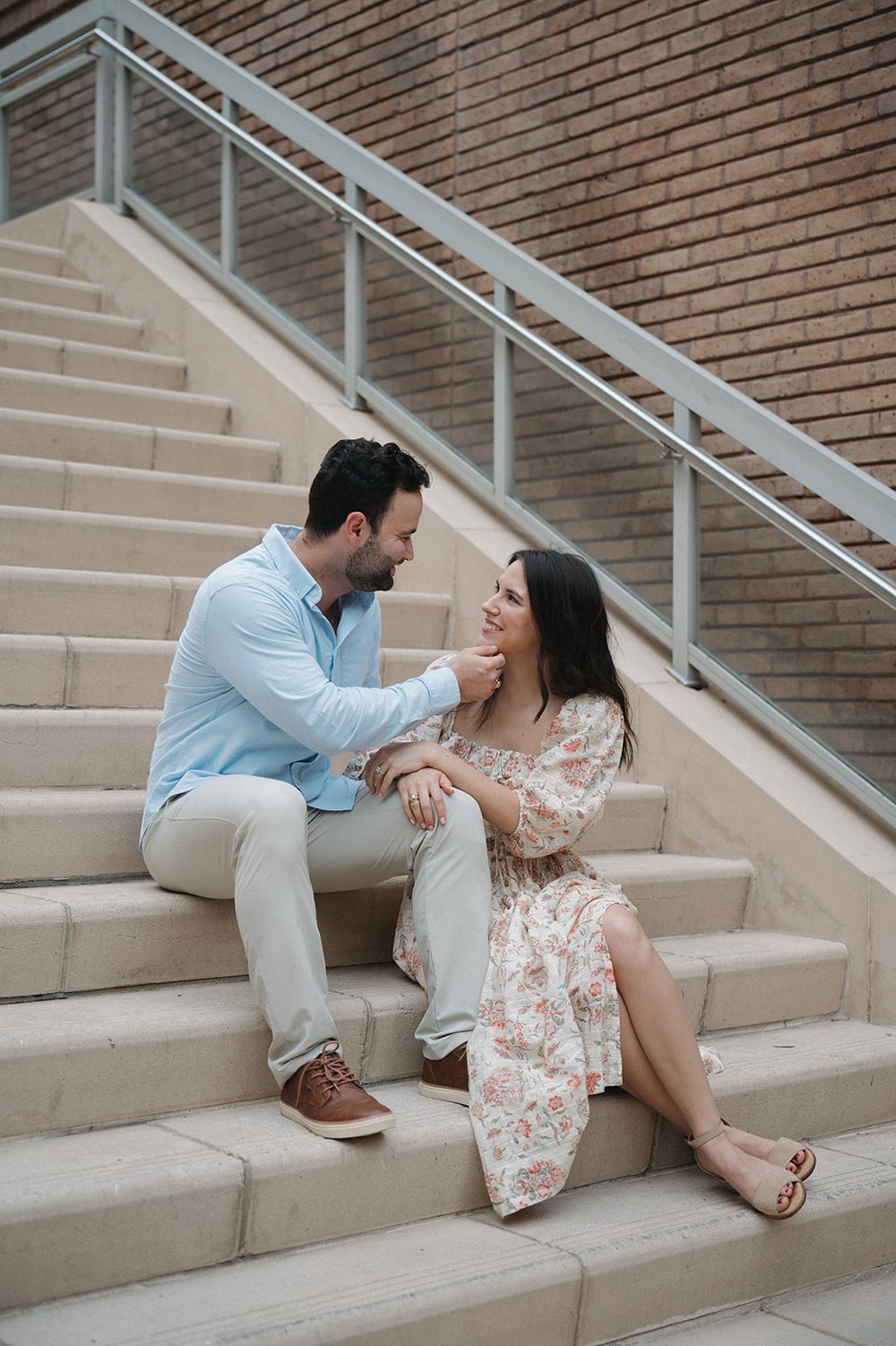 A couple sitting romantically on concrete stairs by a brick wall, she in a floral dress and he in light blue shirt.
