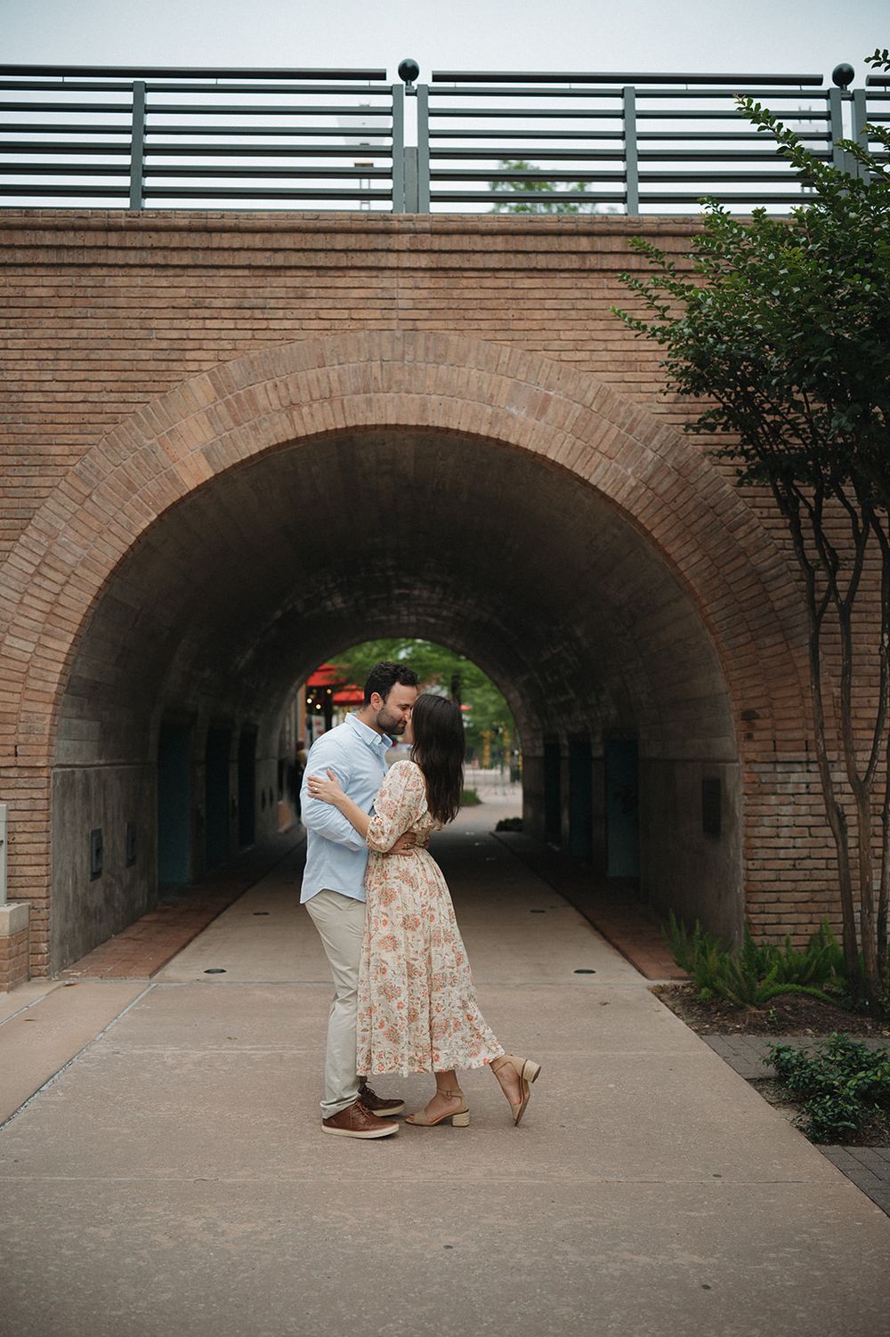 A romantic moment captured under a brick archway tunnel with lush greenery visible in the background.
