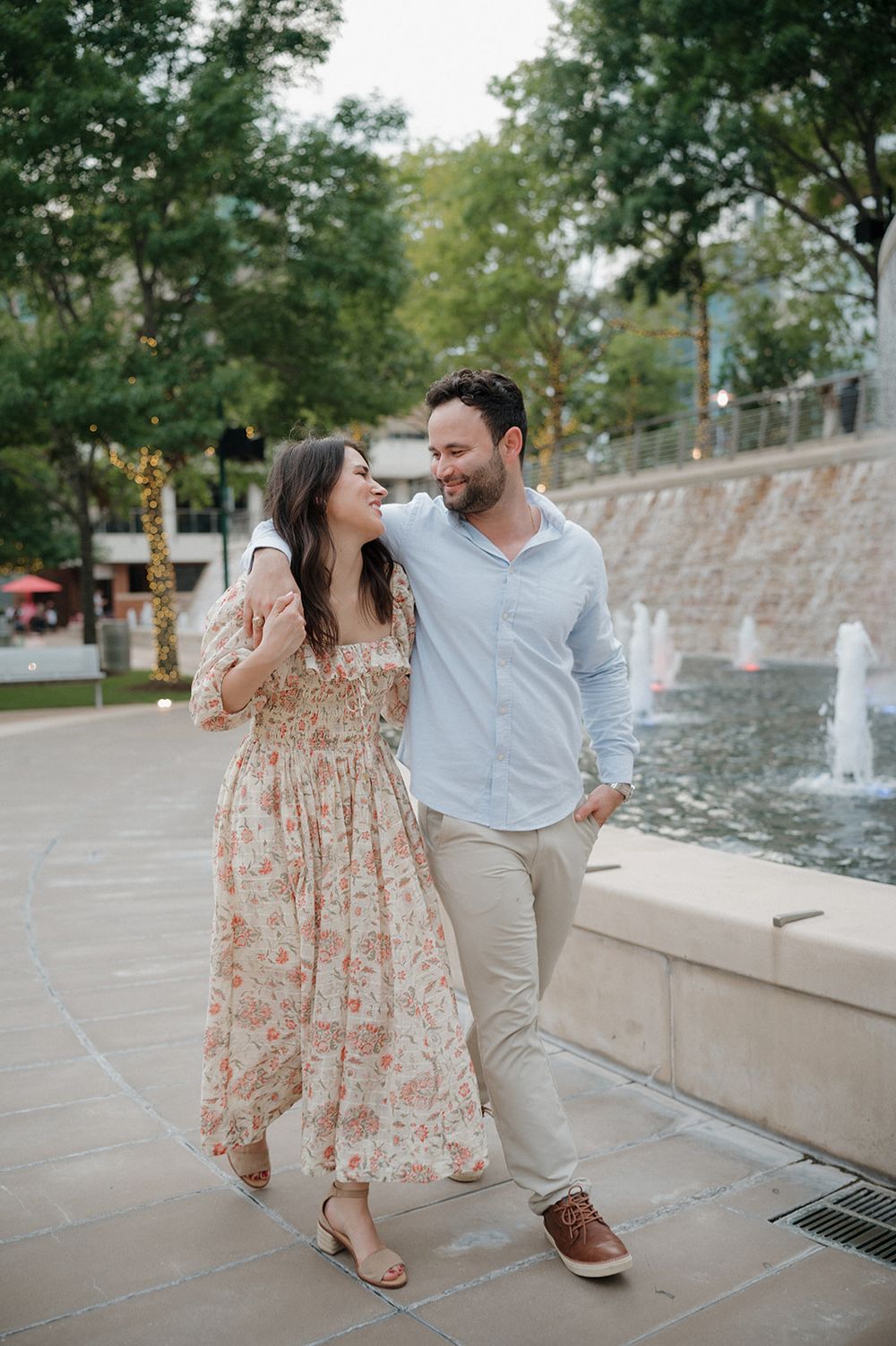 A couple walking and laughing together by a fountain, with the woman wearing a floral dress and man in casual attire.