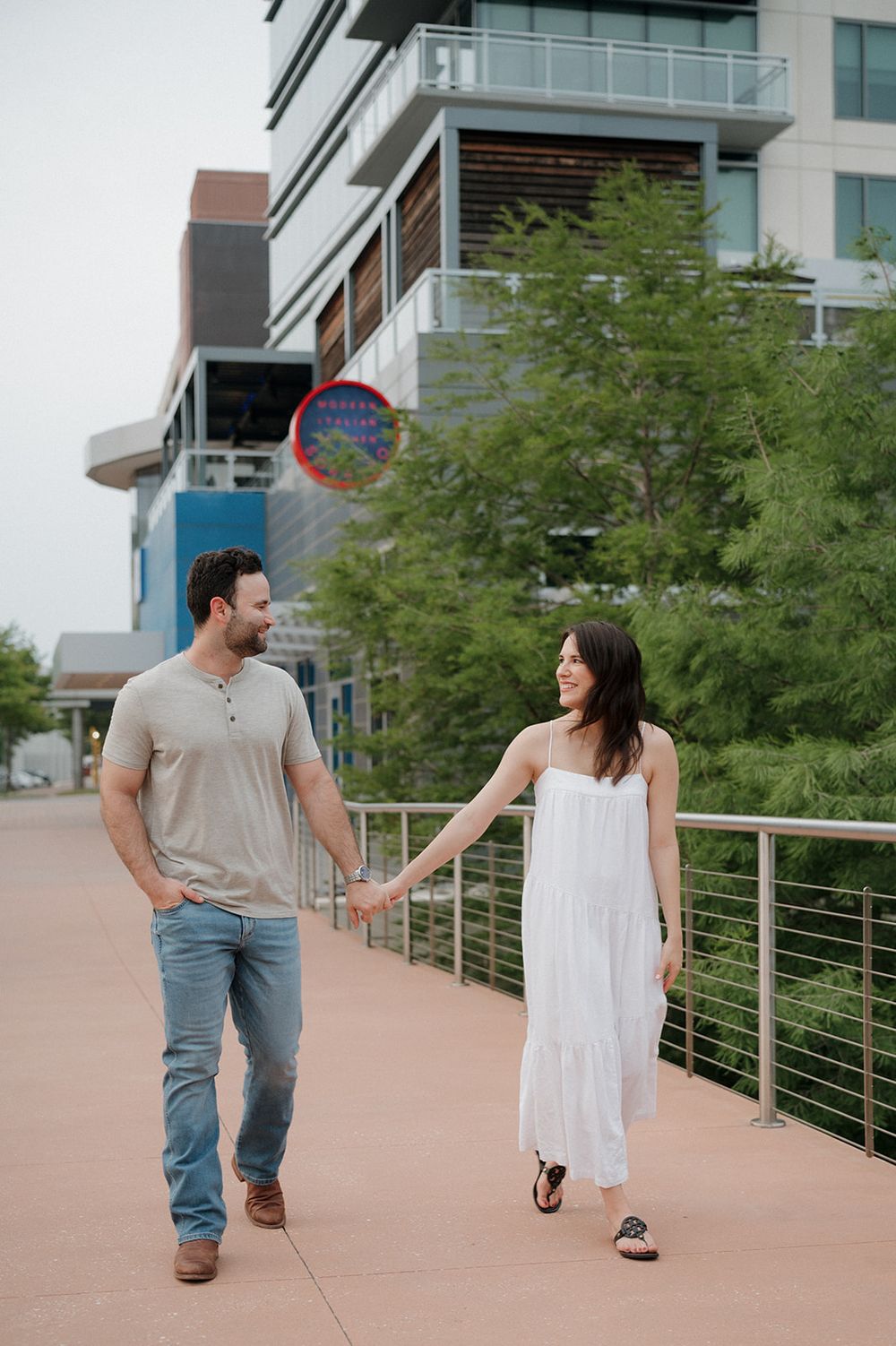 A couple walking together on a modern walkway with contemporary architecture and greenery in the background.